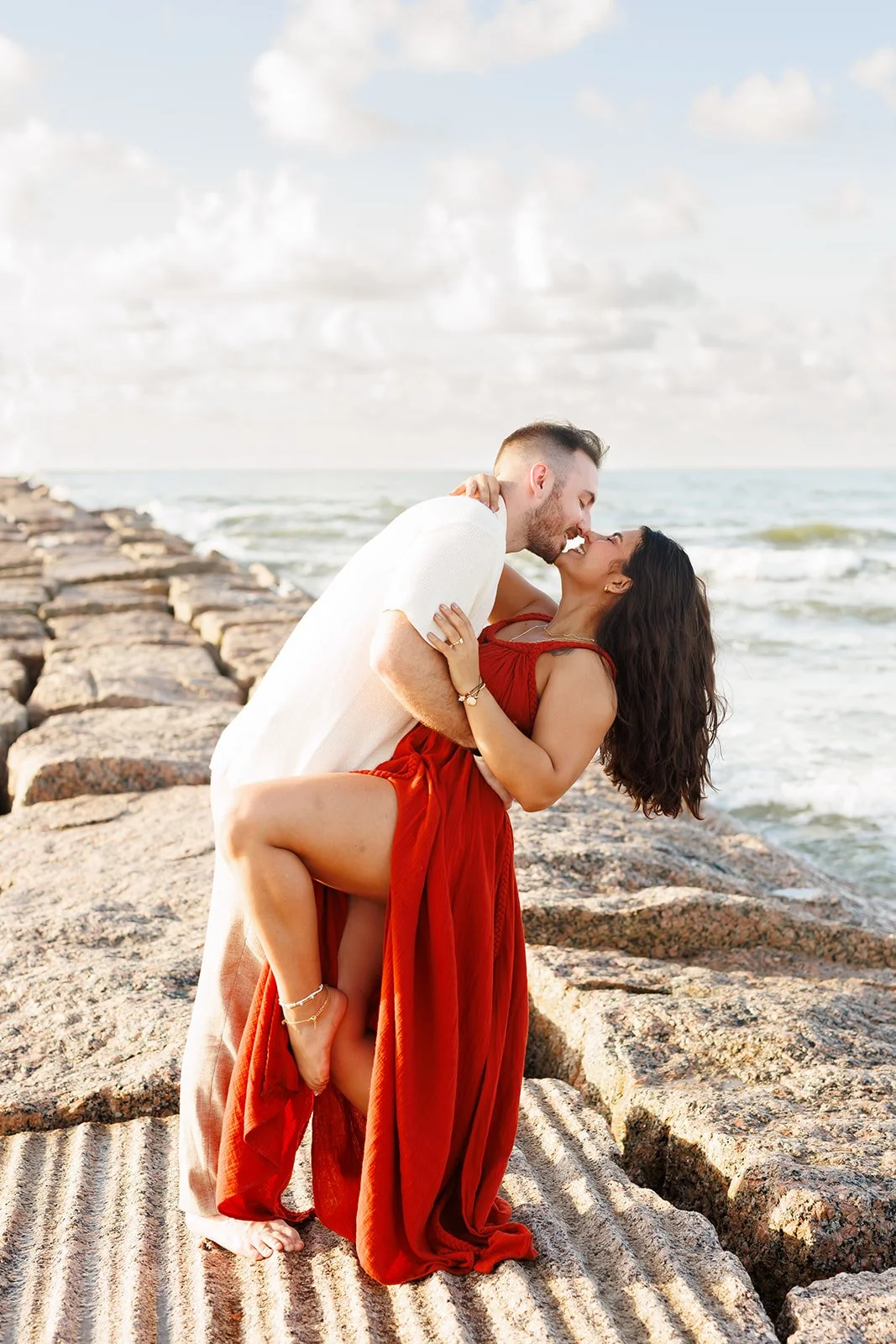 Passionate Galveston beach photo — man lifting and kissing his partner in a red dress with ocean waves behind — Wild Ivy Creative.