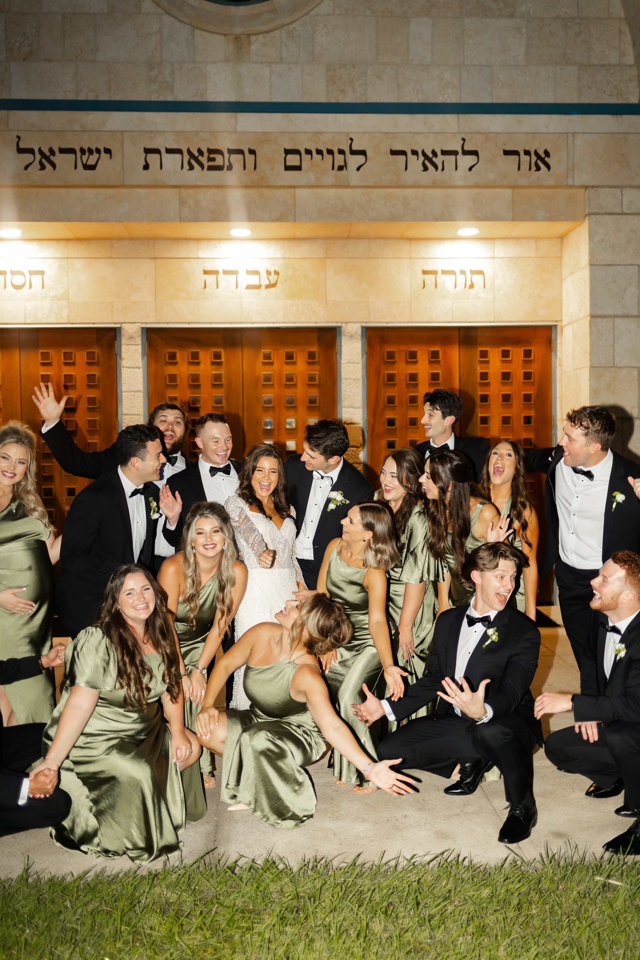 A group of people in formal attire, including tuxedos and dresses, gathered outside a building with Hebrew signs, celebrating and posing for a group photo at a wedding captured by top-rated wedding photographer Wild Ivy Creative Photography