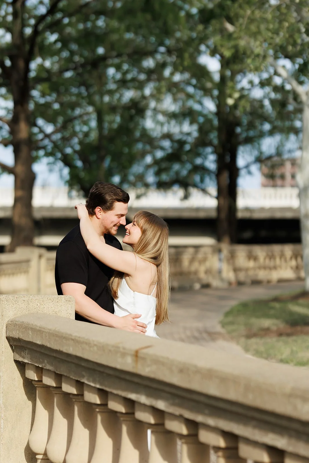Outdoor Downtown Houston engagement photo of a couple embracing near a stone railing and bridge, surrounded by greenery — Wild Ivy Creative.