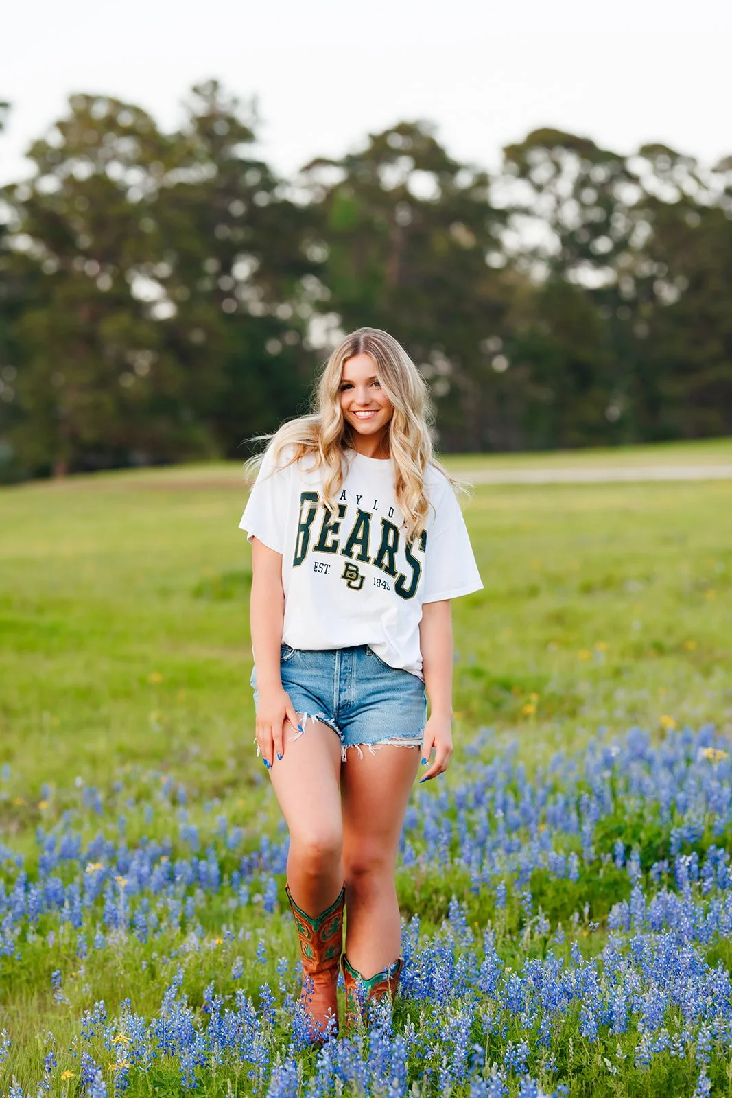 A young woman with long blond hair smiling, standing in a field of blue wildflowers, wearing a white T-shirt with 'BAYLOR BEARS' and denim shorts, with cowboy boots for senior portraits at Blue Jack National captured by Wild Ivy Creative
