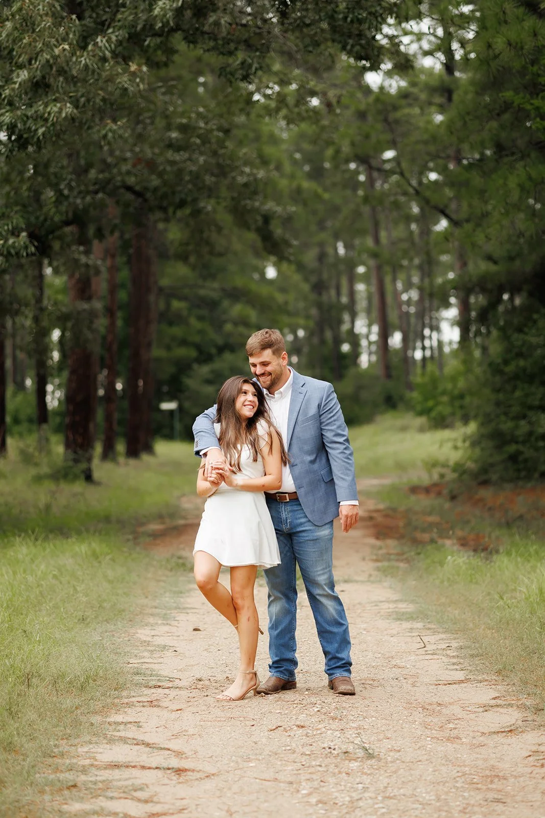 A young woman and a man walking together on a dirt path in a wooded area, smiling and holding hands. The woman has long hair and is wearing a white dress, while the man has short hair and is wearing a light blue blazer and jeans.