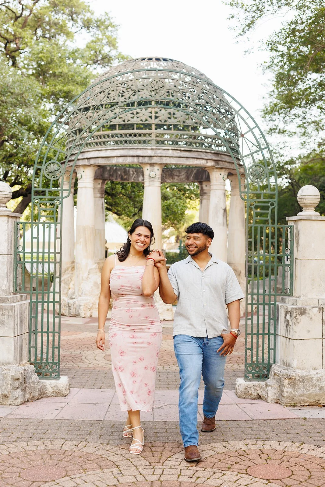 Romantic park proposal session in Houston with a couple walking hand-in-hand through a decorative gate — by Wild Ivy Creative.