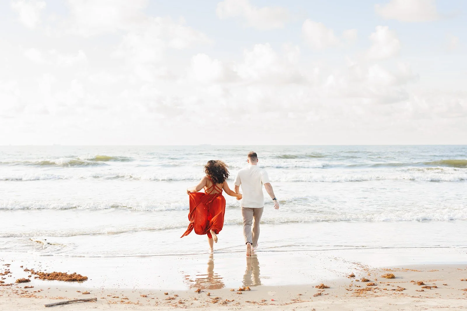 Playful Galveston beach engagement photo — couple walking hand-in-hand into the surf beneath a cloudy sky — Wild Ivy Creative.
