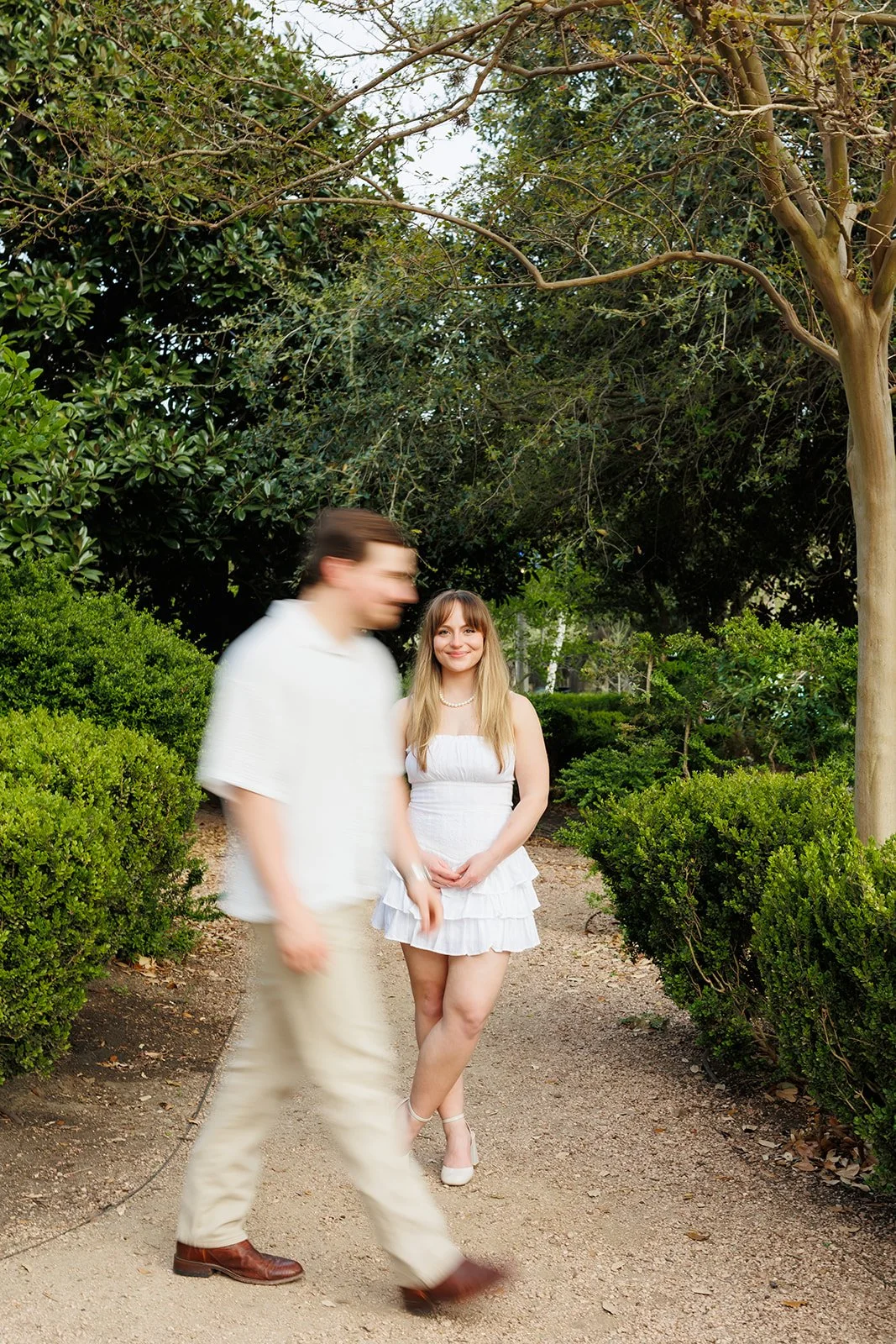 Artistic Houston engagement photo — woman in white smiles on wooded path while her partner passes in soft motion blur — Wild Ivy Creative.