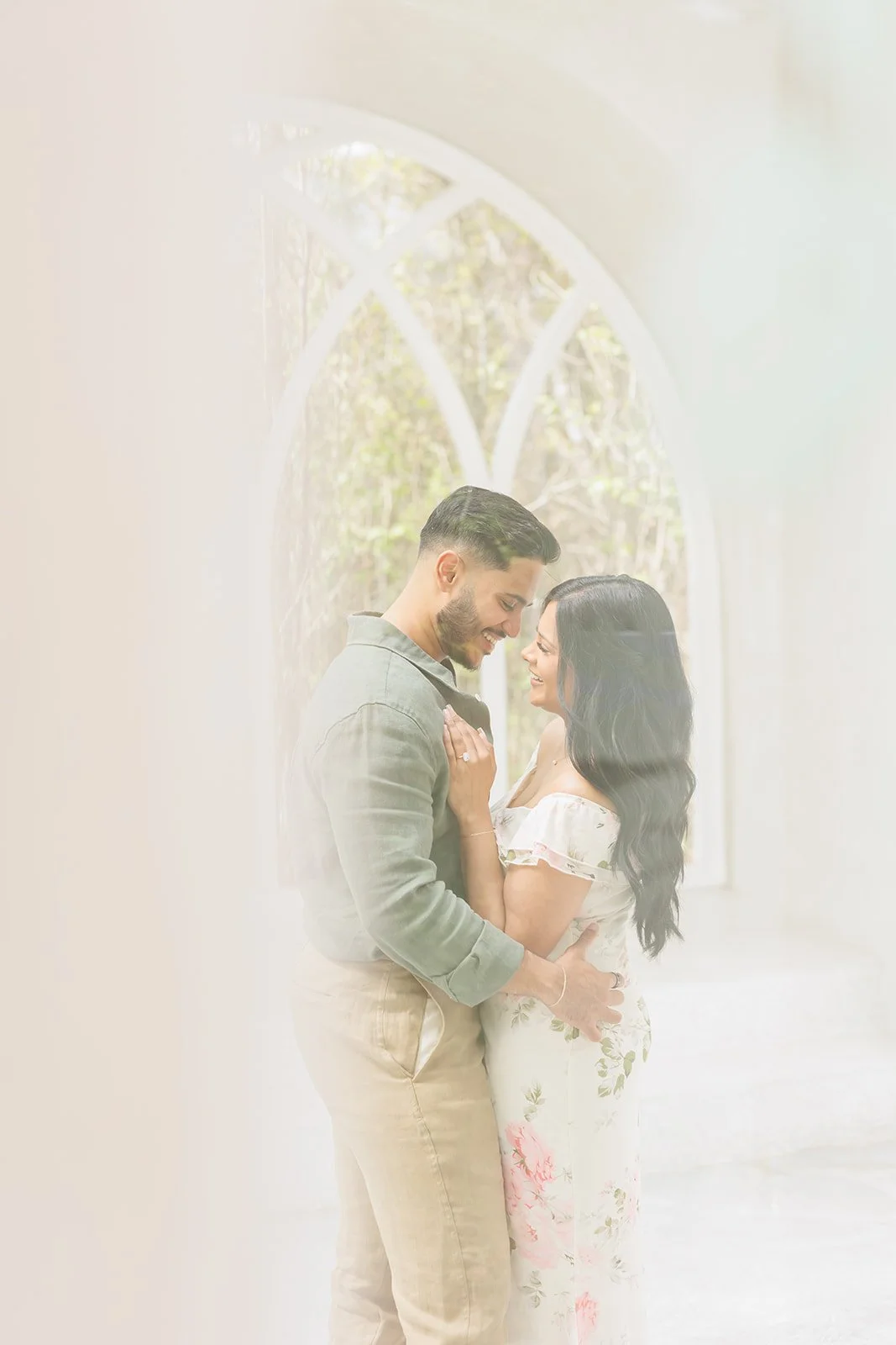 Soft and romantic engagement photo of a couple embracing by a large sunlit window during a Houston photography session.