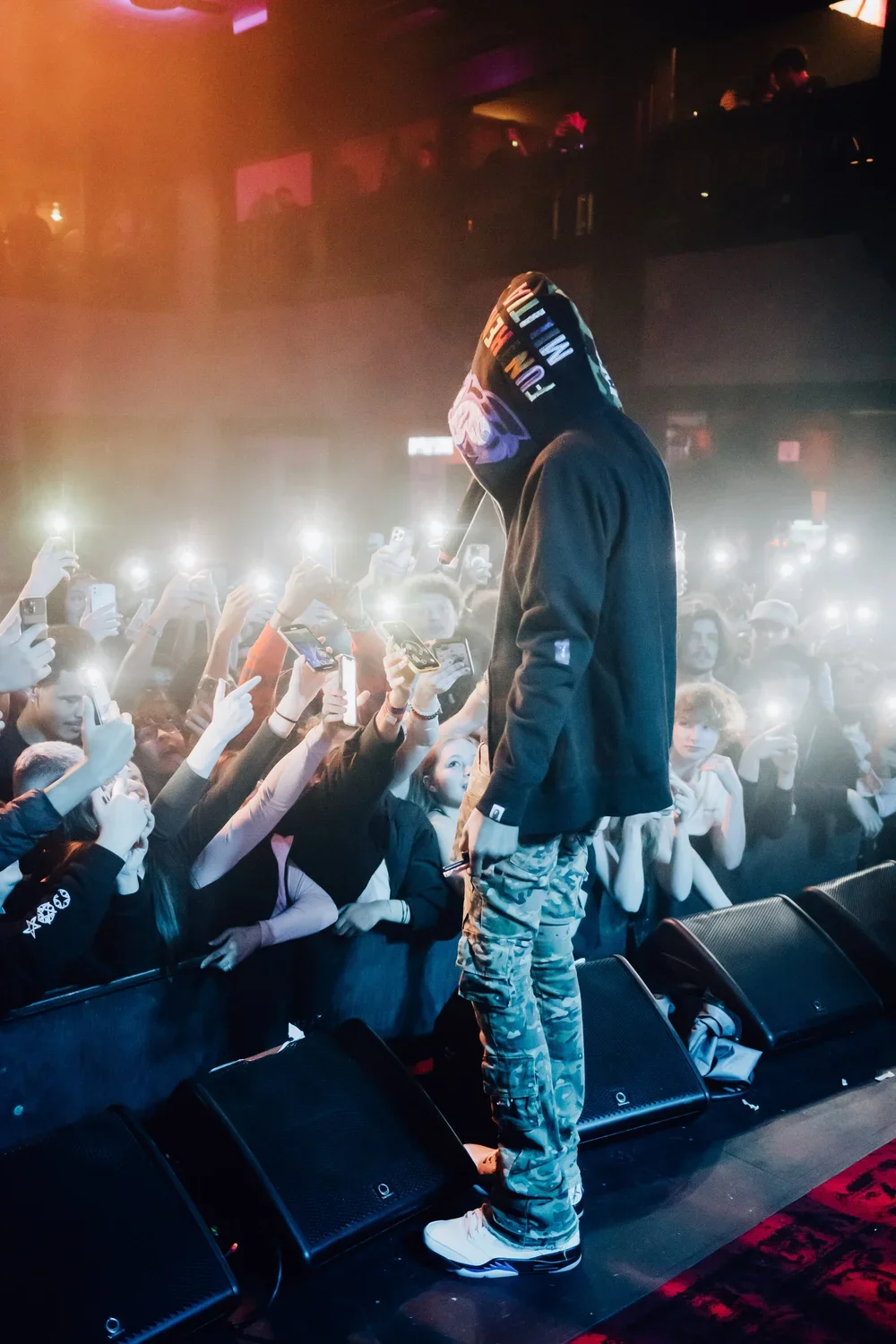 Performer on stage wearing a hoodie with a colorful design, facing a crowded audience at a concert or nightclub.