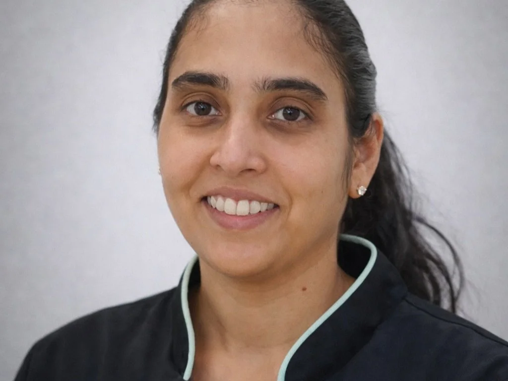 Close-up of a smiling woman with dark hair tied back, wearing a black uniform with light-colored piping, and earrings, against a plain background.