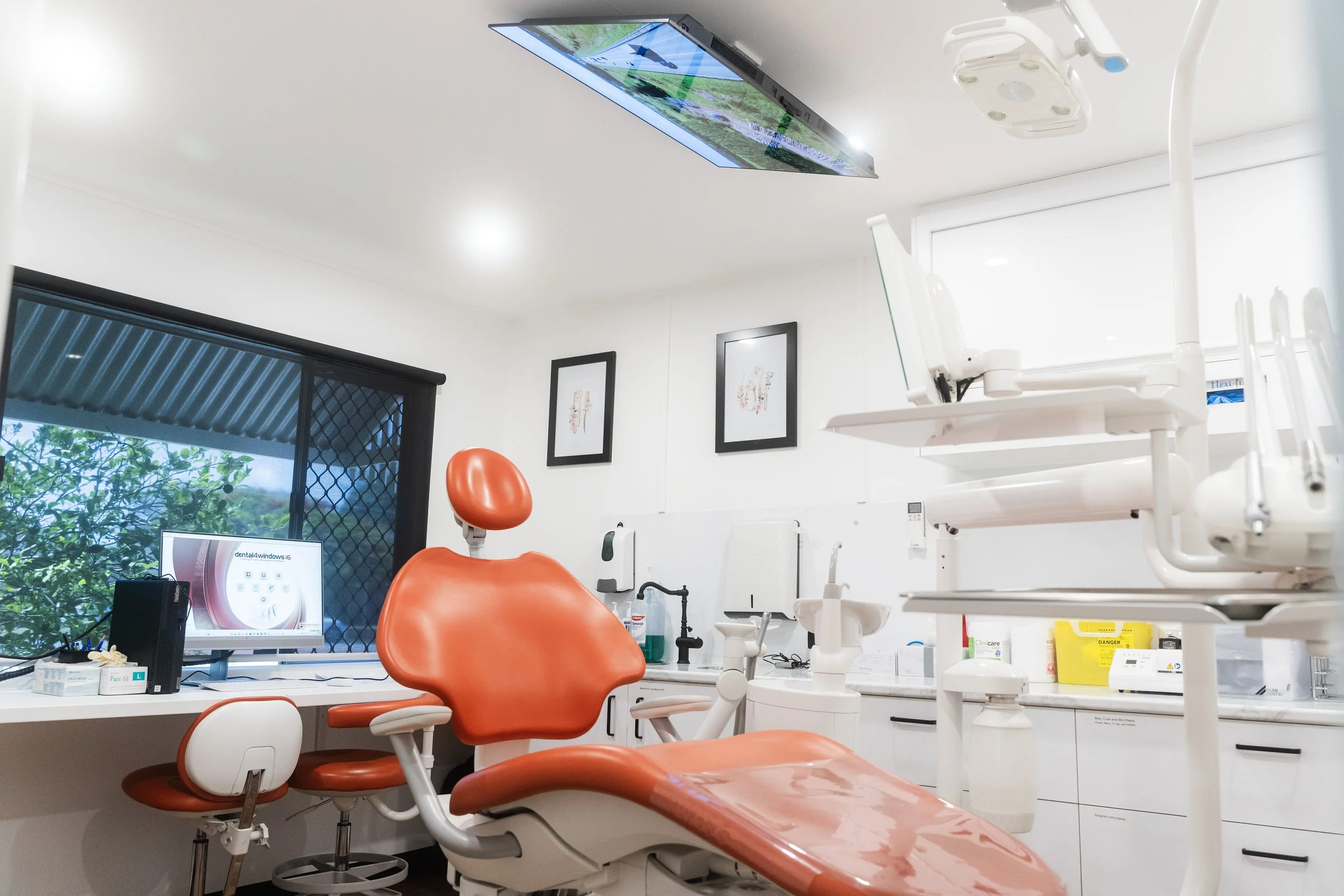 Dental examination room with orange dental chair, dental equipment, window with view of trees, and framed artwork on the white wall.