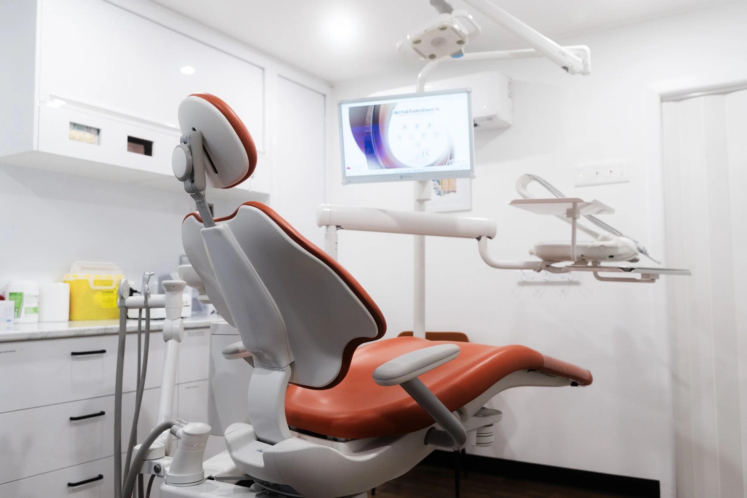 An empty dental clinic with a dental chair, overhead monitor, medical equipment, and white walls.