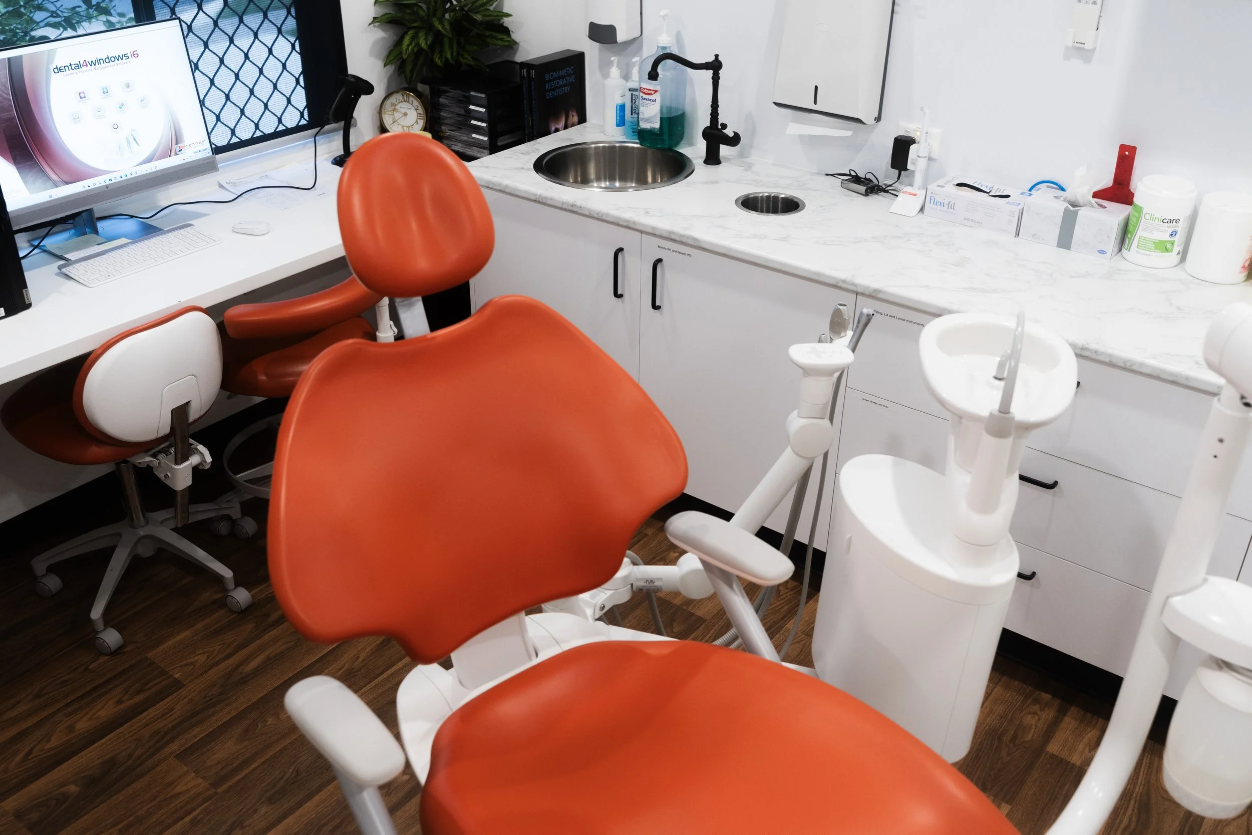 Dental exam room with orange dental chair, white dentist stool, computer monitor, dental tools, and supplies on white countertops.