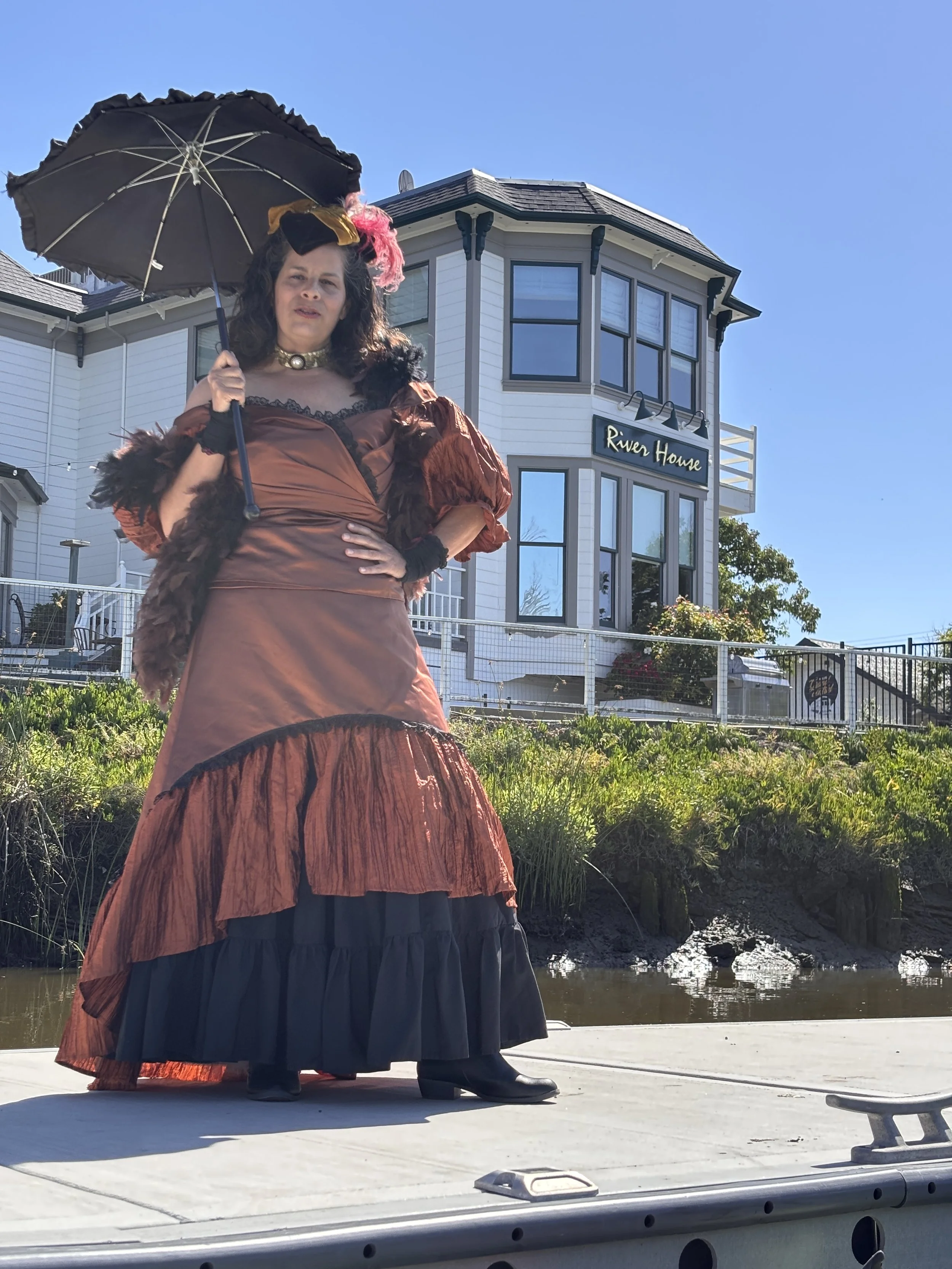 Woman in turn of the century red frilly dress with sun umbrella stands before white Victorian home on a dock