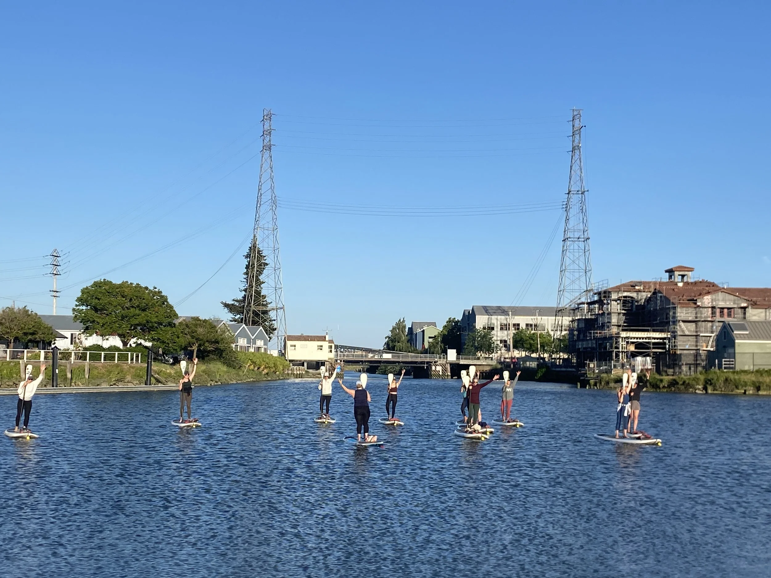 Stand up Paddle Board Yoga in the Petaluma River Turning Basin. D Street bridge and power poles are in the background. (2025)