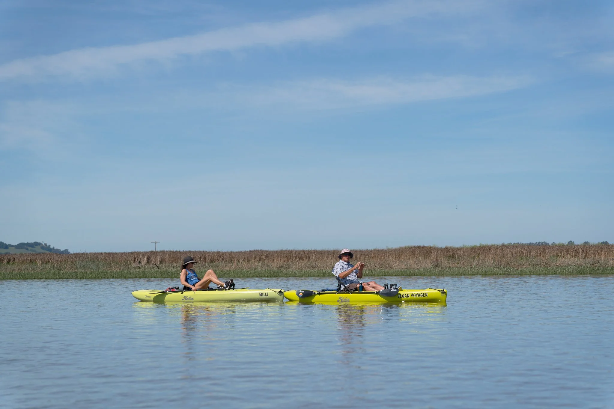 Two people in pedal kayaks float in a landscape and skyscape of blue and green. The reeds behind them are brown and green, and the huge sky is blue, streaked with clouds. They both wear bucket hats and their kayaks are lime green.
