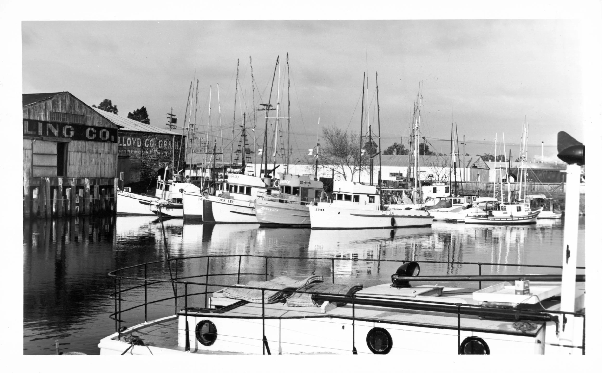 Pleasure boats crowd a dock. Era: 1960s. In the background is an old wooden warehouse reading Golden Eagle Milling Company. The water is calm and reflects the yachts.