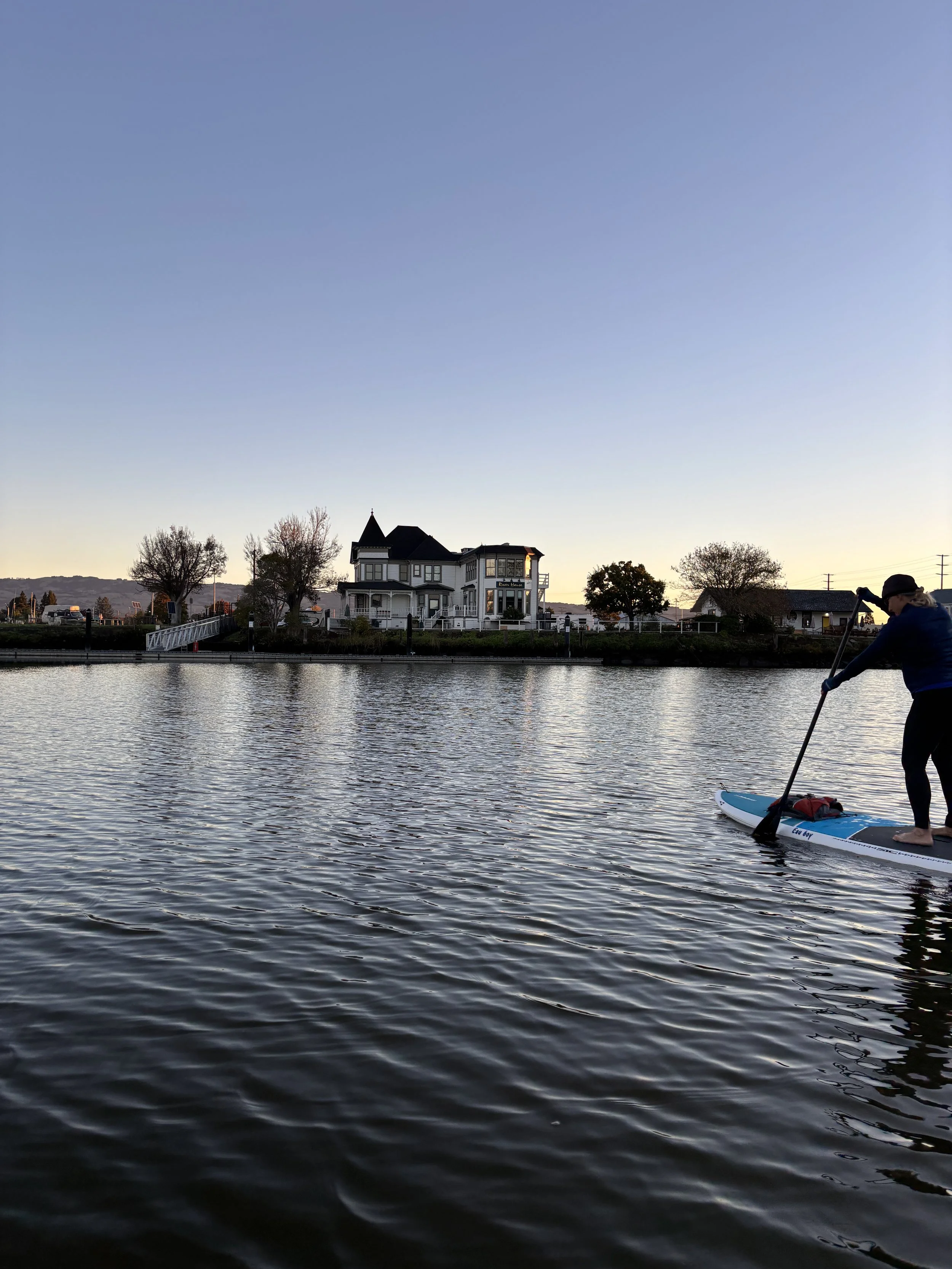 Woman on a paddle board paddles toward a Victorian house  at dawn on calm water