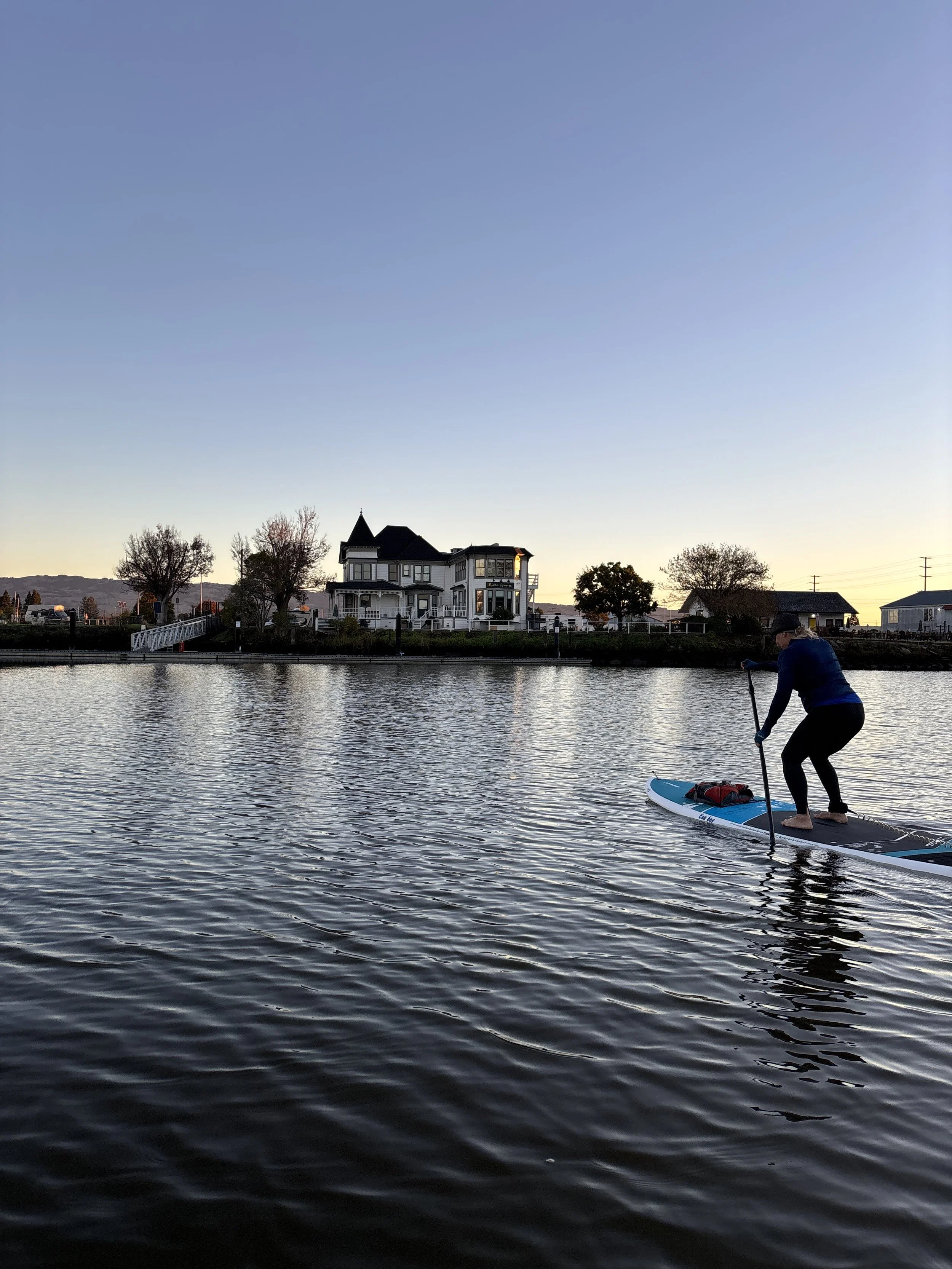 A woman paddles on a river at dawn. The water is rippling, and a white Victorian home is visible in the background against the brightening sky.