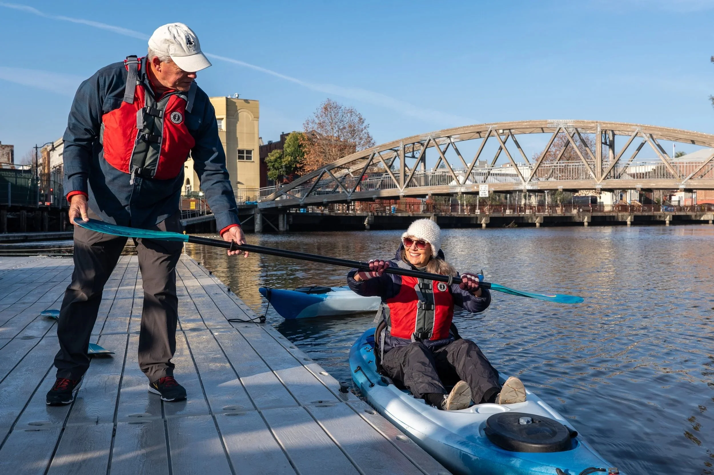 Man on dock hands a paddle to a woman in a kayak, who is wearing a white beanie hat and smiling. Both people are wearing personal flotation devices and there is a wooden footbridge in the background. 