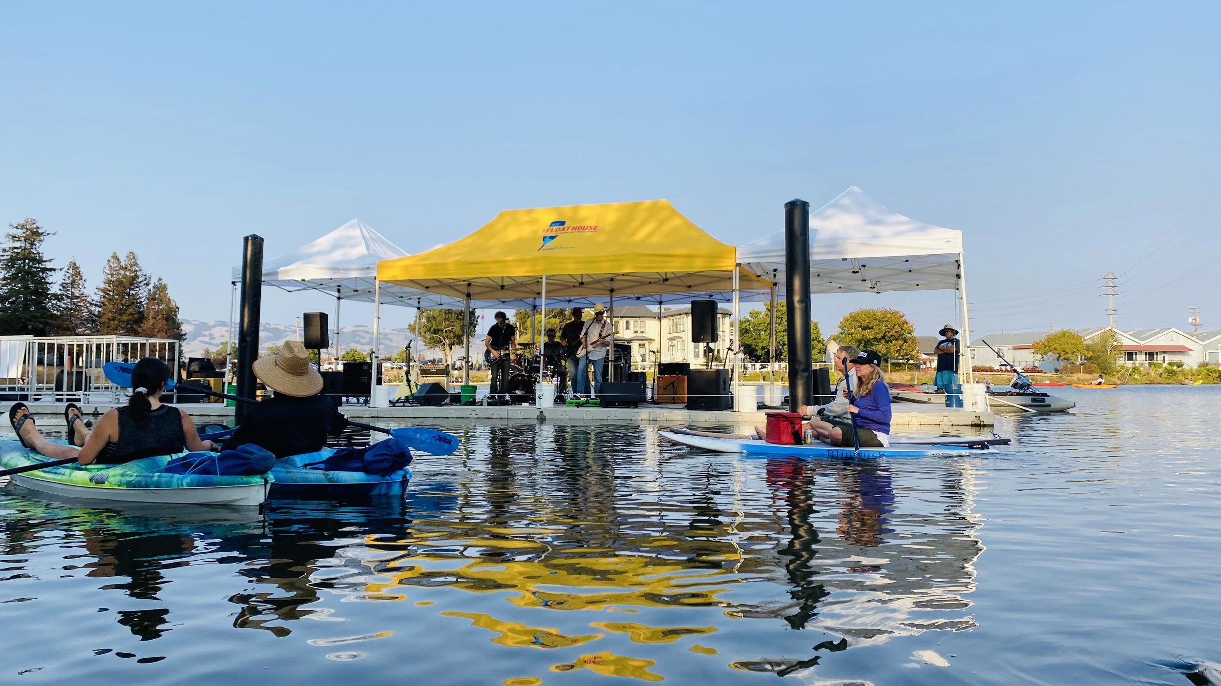 A yellow tent on a white dock. People in kayaks with paddles sit on shimmering water reflecting the tent and the band members. Four musicians play guitar, bass, keyboards and drums. The sky is blue.