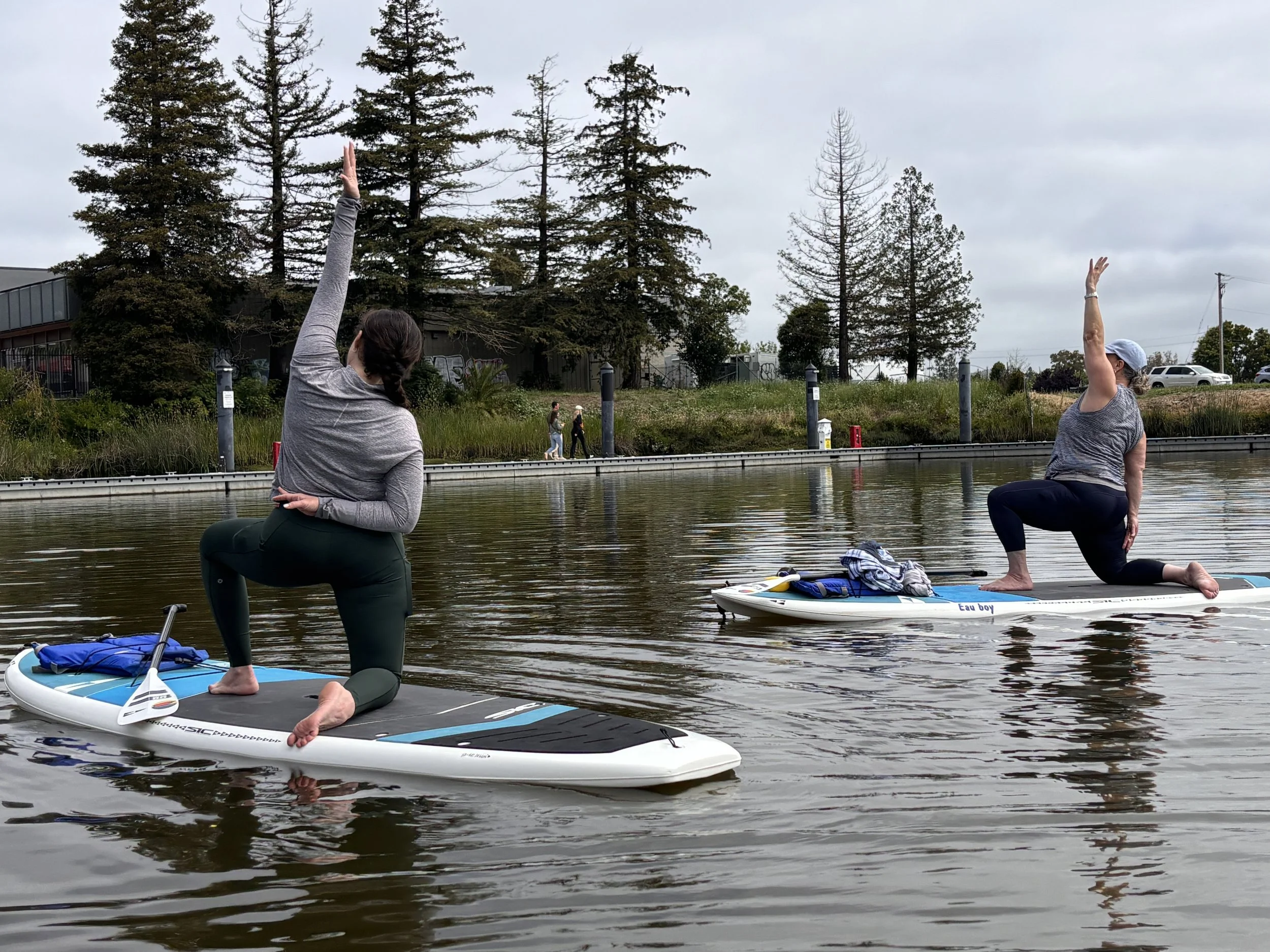 Two women do reverse warrior pose on their knees on paddleboards on a body of water. There are redwood trees and a dock in the background. The sky is cloudy.