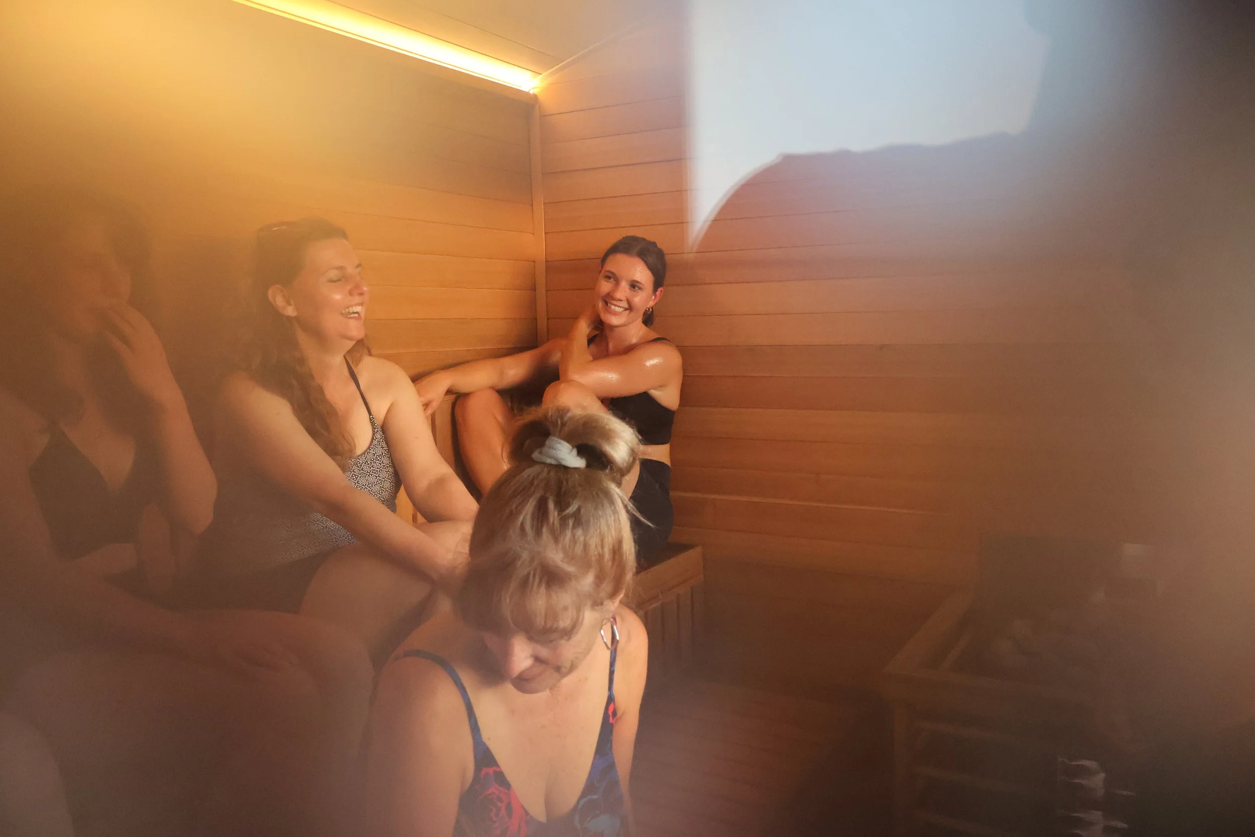 Four women laugh inside of a wooden sauna room. Sunlight streams through a picture window. They are sweating.