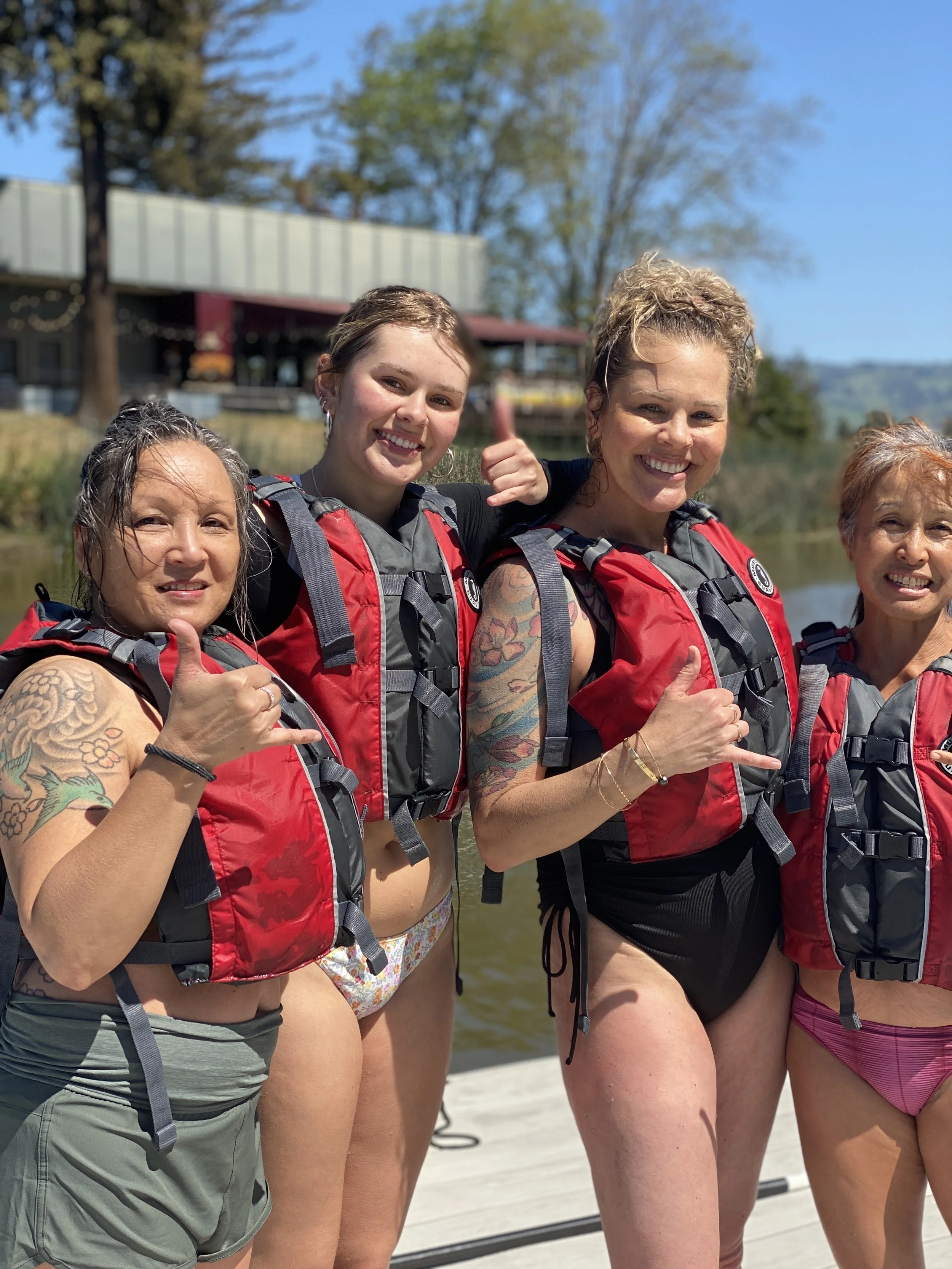 four women in red personal flotation devices holding their hands in the Hawaiian aloha way, smile in front of a body of water.