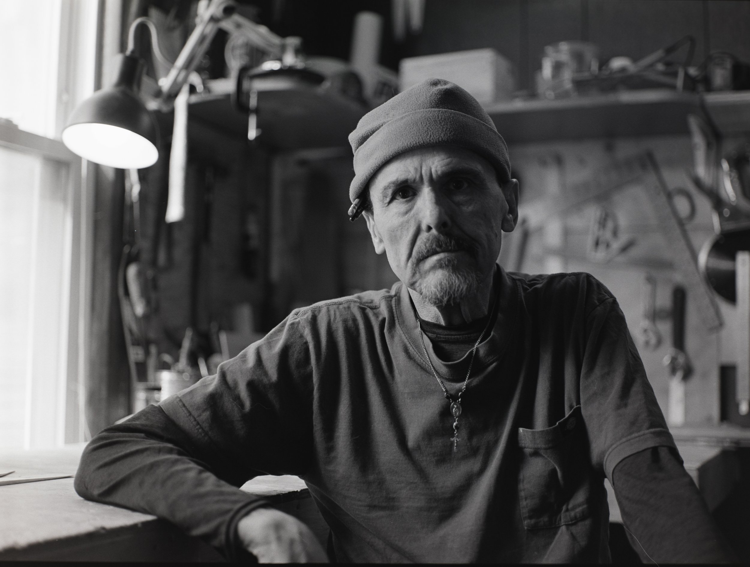 A man with a beard and mustache wearing a beanie hat and a necklace with a cross pendant, sitting in a workshop with tools on a wall and a lit desk lamp in the background.