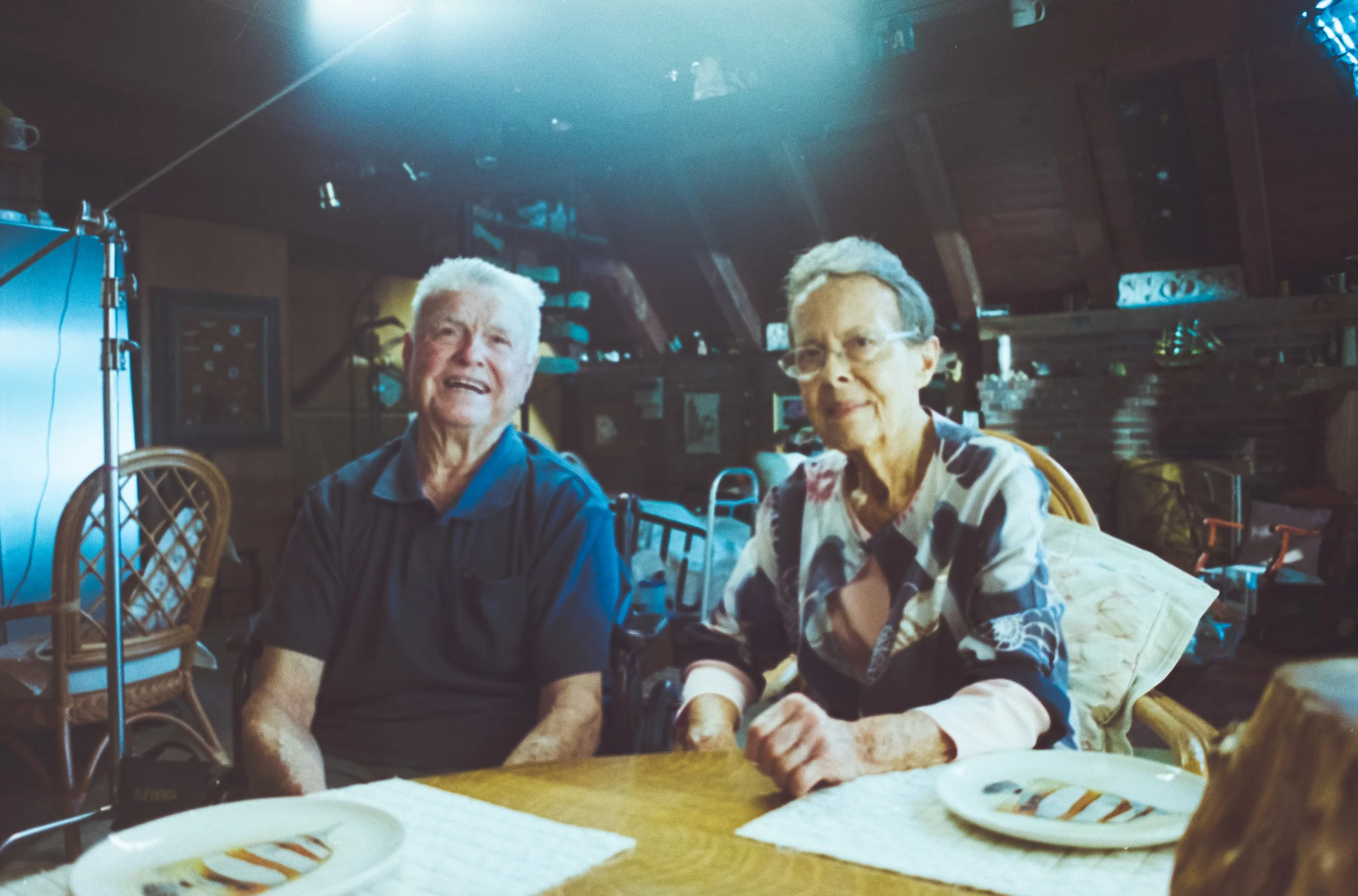Two elderly people sitting at a table in a cozy, wooden-paneled room with two empty plates in front of them. The man is wearing a dark shirt and the woman is wearing a floral blouse. There are chairs and shelves in the background.
