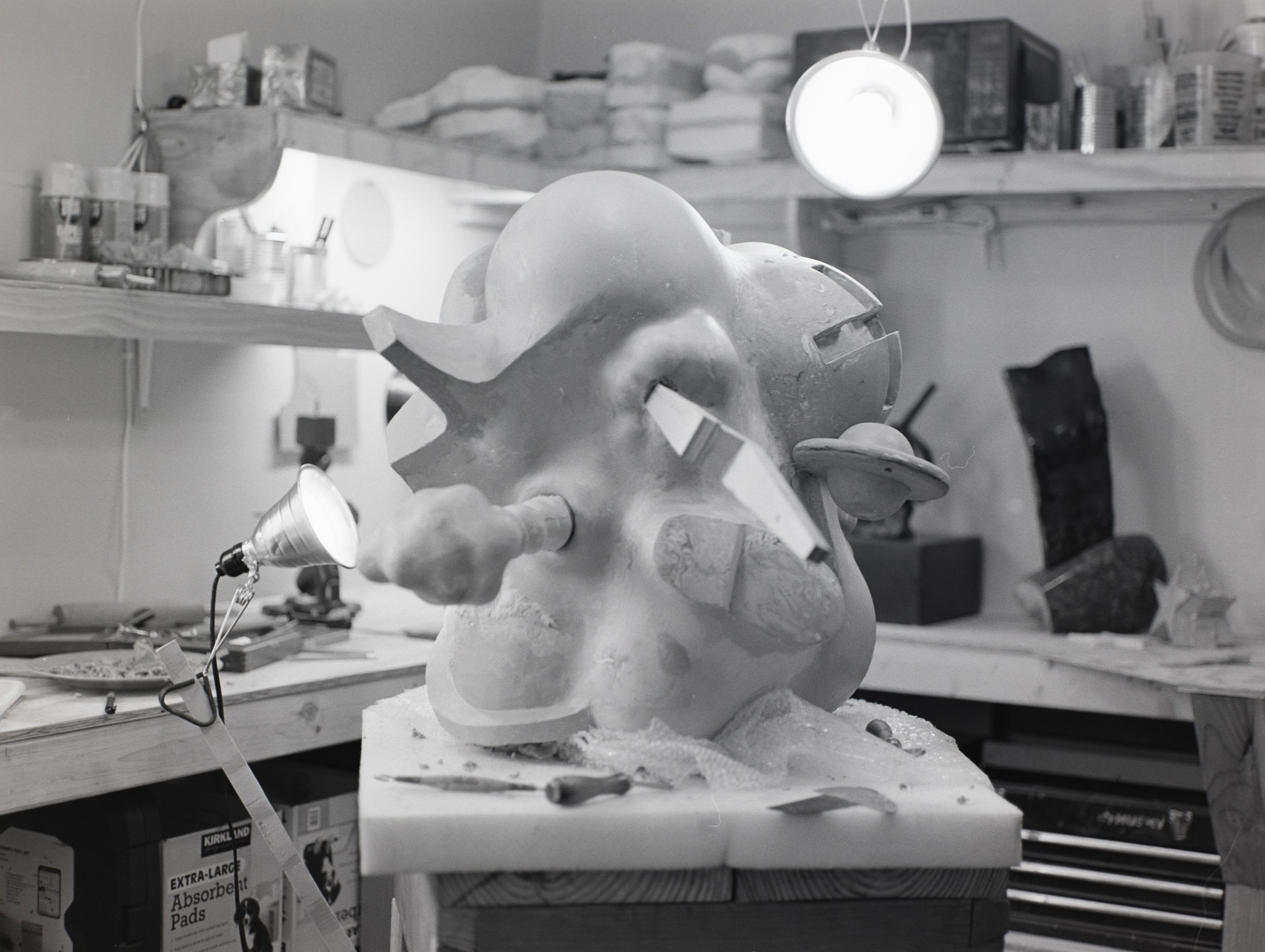 A workspace with a sculpture of a humanoid face in progress, surrounded by tools and materials. There is a lamp illuminating the sculpture, with shelves holding various supplies in the background.