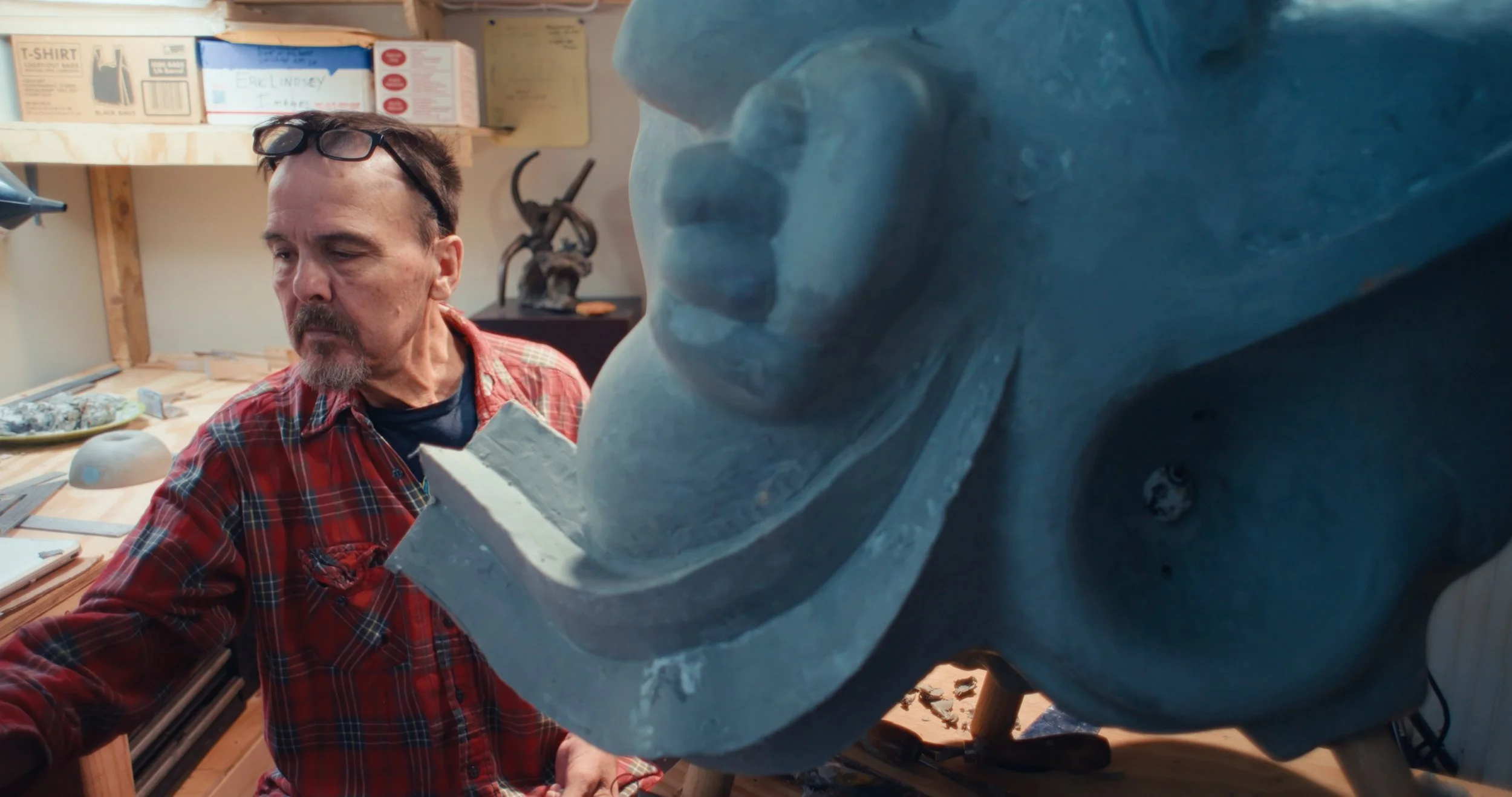 An artist in a workshop working on a large sculpture of a wolf's face, with various tools and materials around.