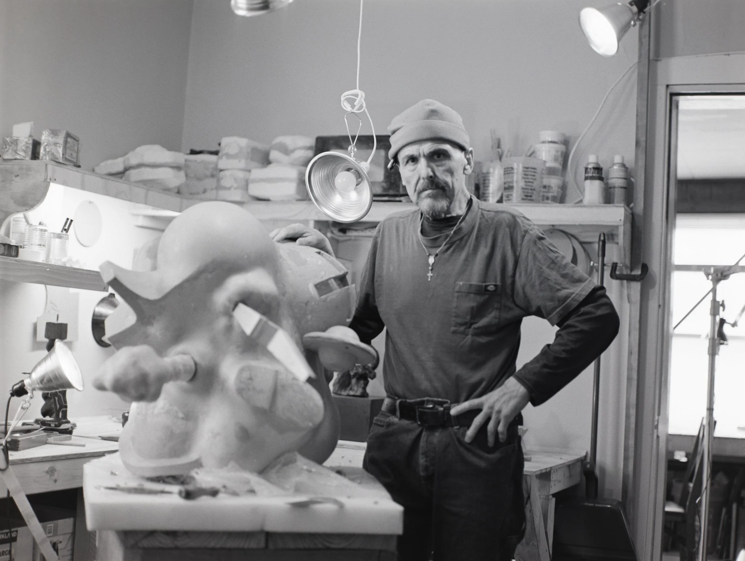 A man with a beard, wearing a beanie, with a serious expression, standing in a workshop. He has a necklace with a cross. There is a sculpture in progress on the workbench in front of him. The workshop has shelves with supplies and a door with a windo