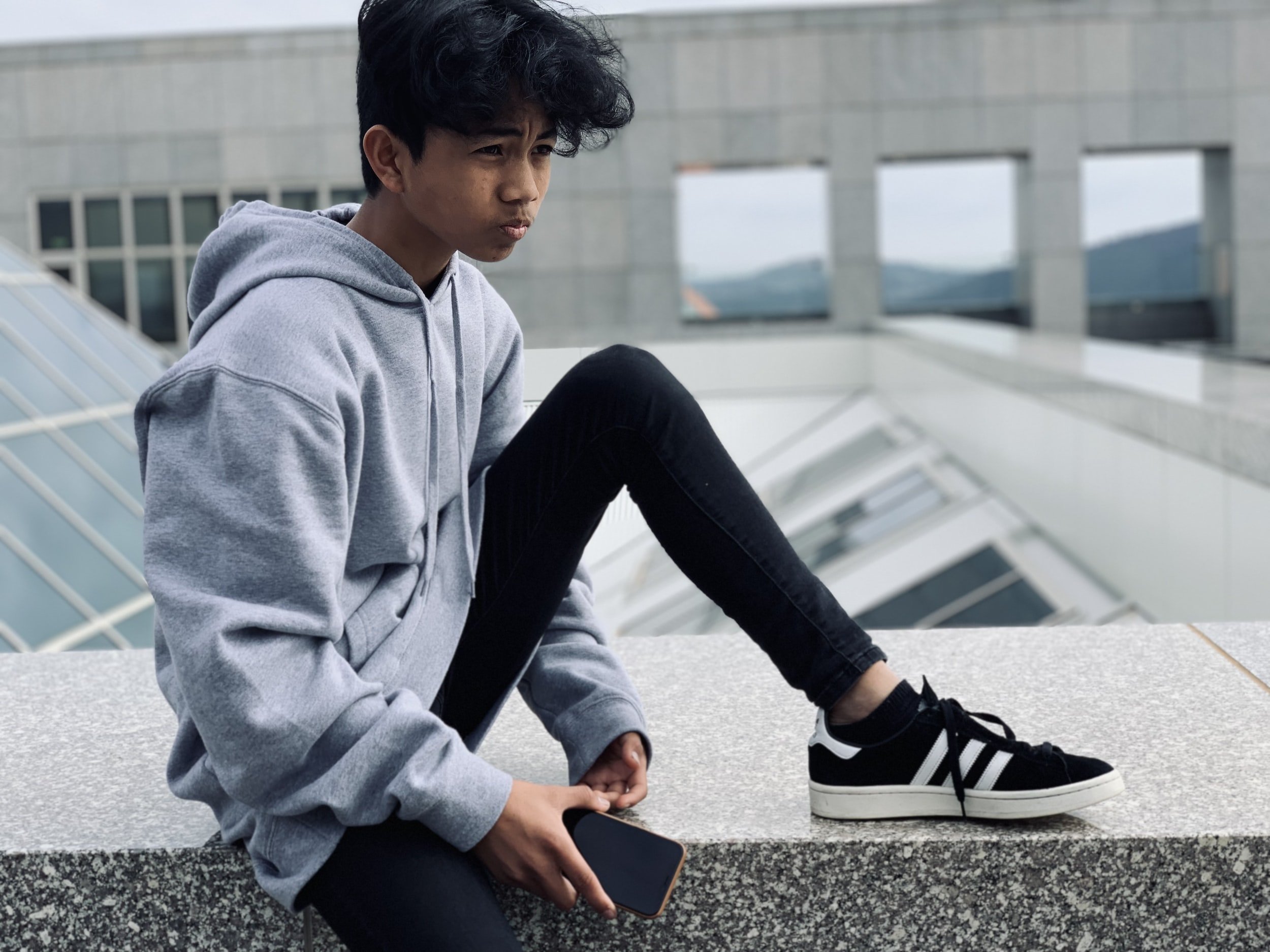 Young man sitting on a concrete ledge, wearing a gray hoodie, black skinny jeans, and black sneakers with white stripes, holding a smartphone, with modern building architecture and windows in the background.