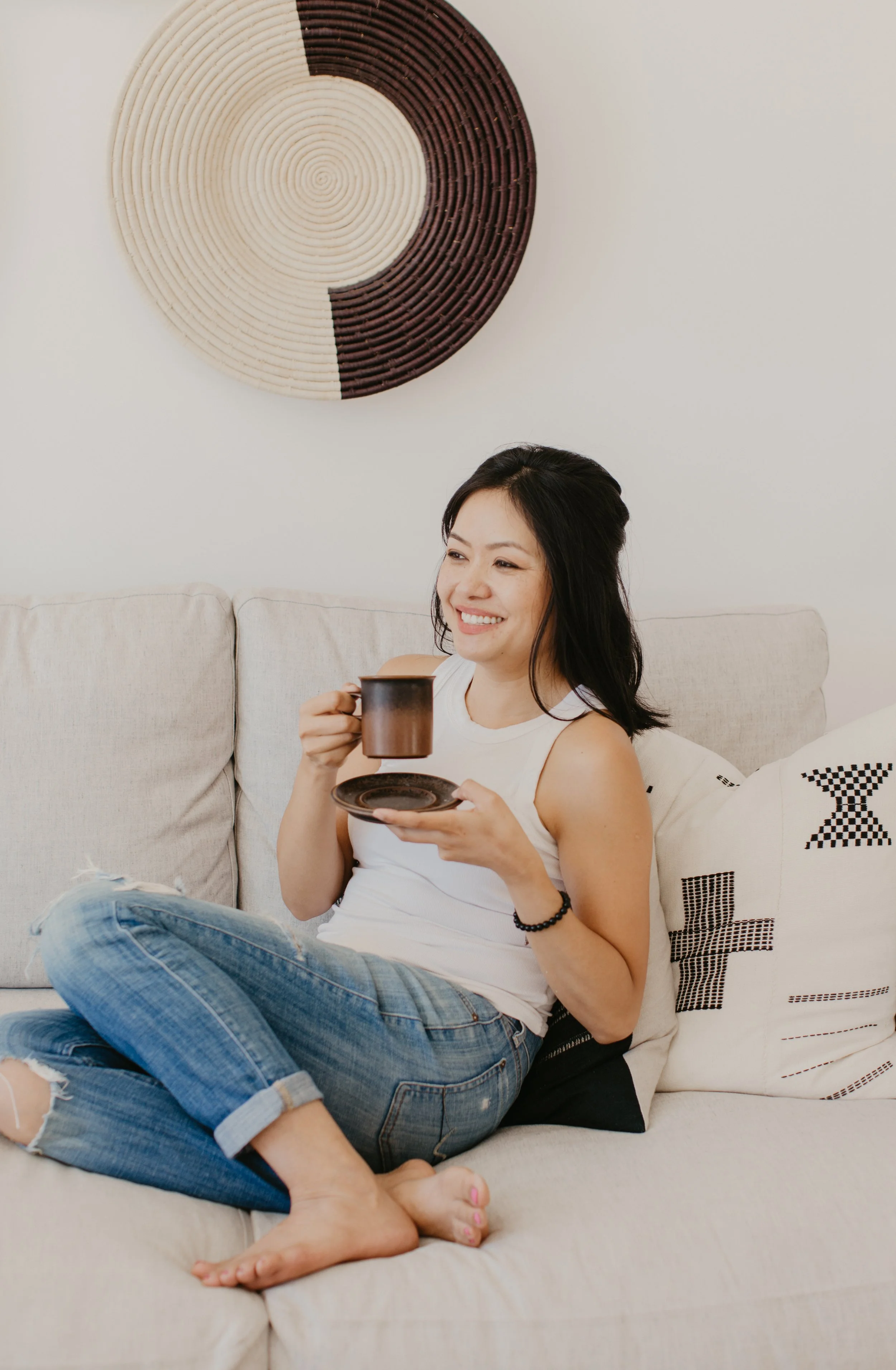 A woman smiling and sitting on a beige couch, holding a coffee mug and a saucer, with her legs crossed. She is wearing a white sleeveless top and ripped jeans. Behind her is a decorative pillow and a piece of circular wall art with contrasting segments in brown and cream.