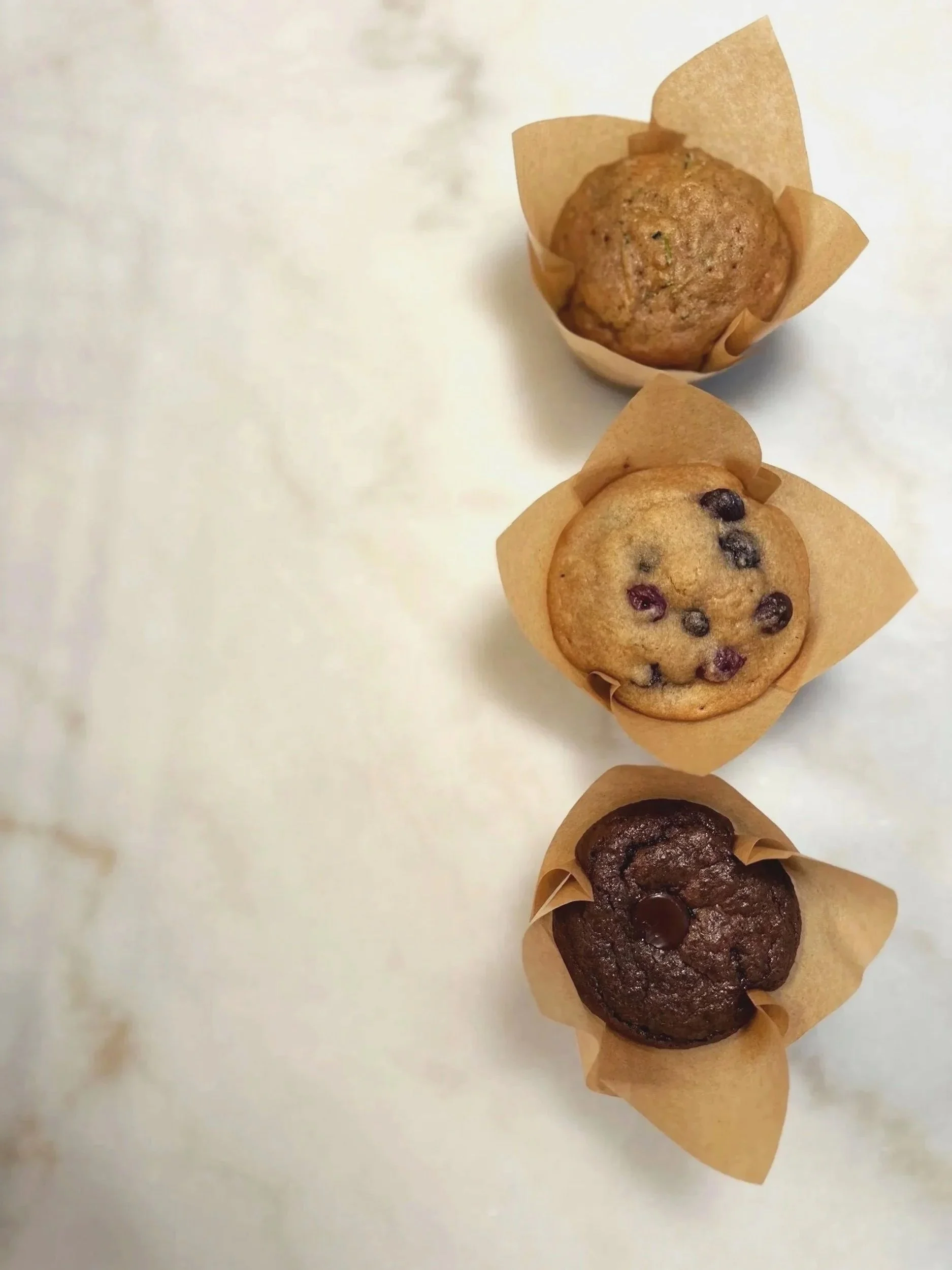 Four muffins with different flavors and toppings, each in a paper wrapper, arranged in a slight diagonal on a white surface.
