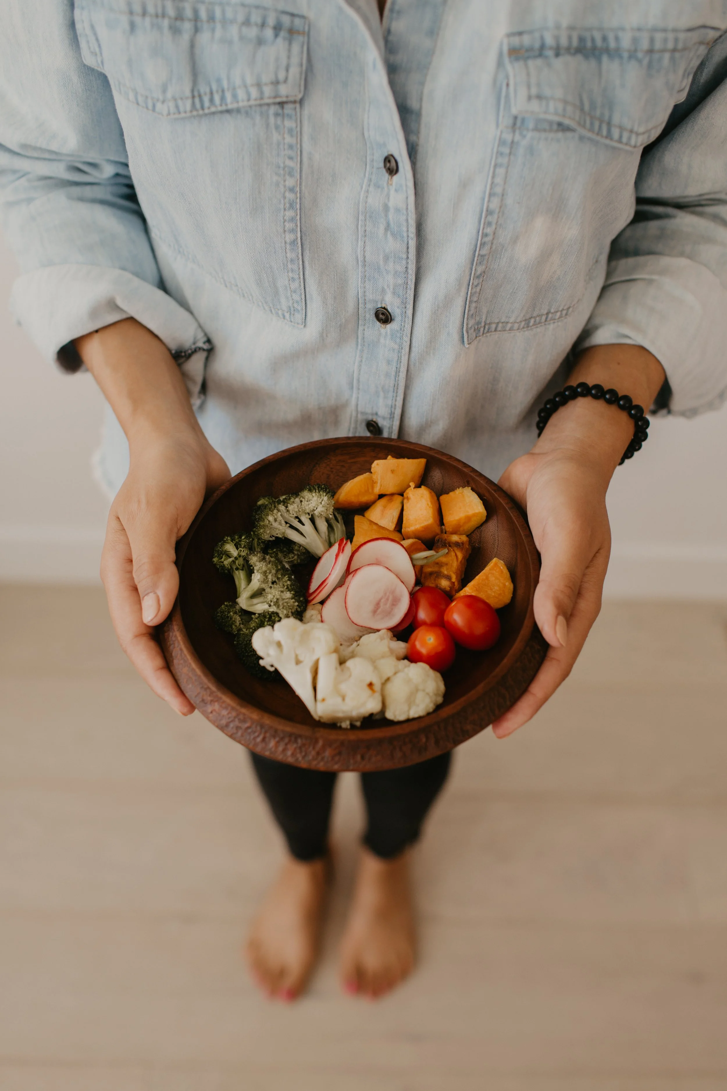 Person holding a wooden bowl of fresh vegetables, including cherry tomatoes, cauliflower, radishes, broccoli, and sliced root vegetables, wearing a light denim shirt with rolled-up sleeves and a black beaded bracelet.