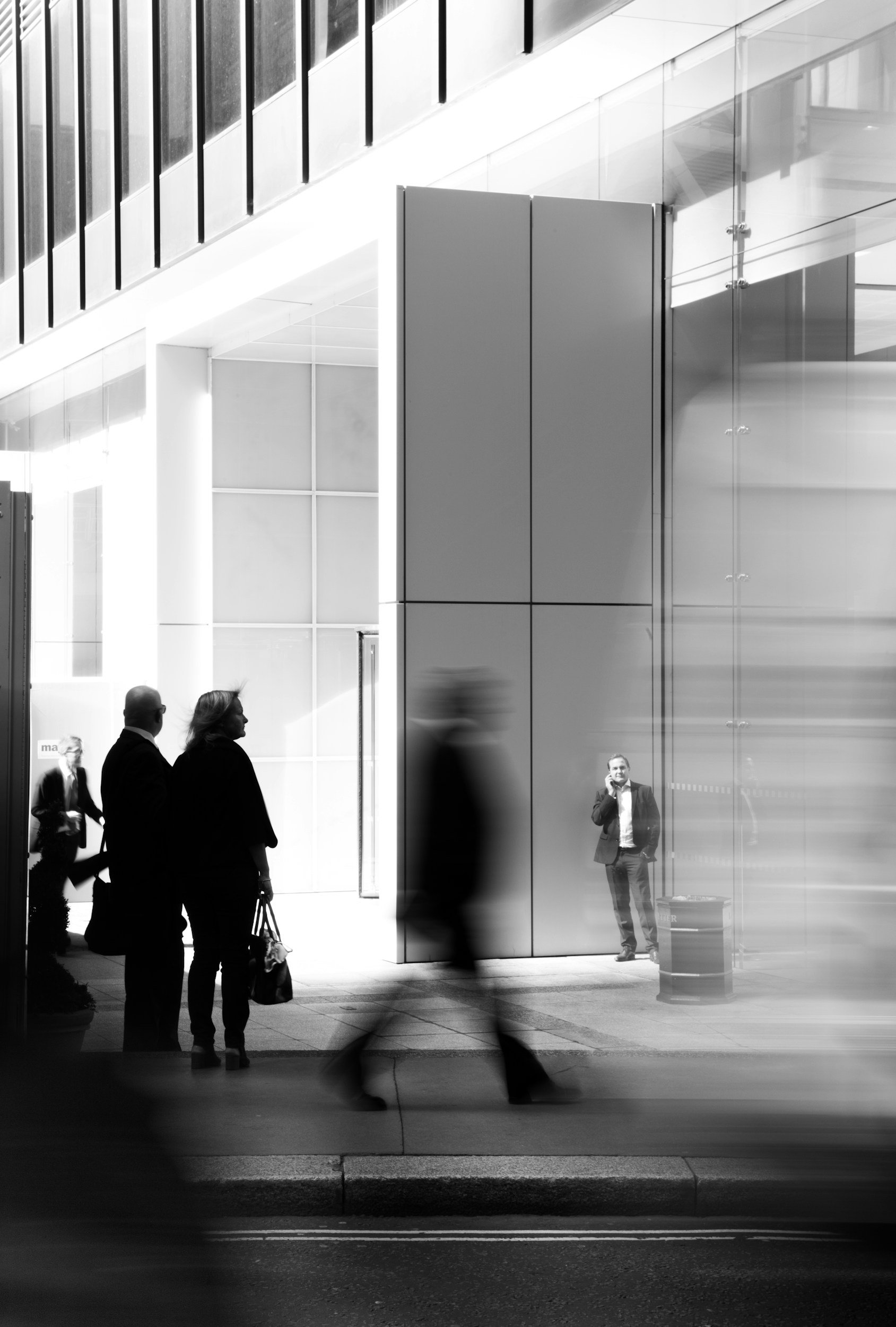Black and white image of a modern office or train station with several blurred people moving past and a man talking on a phone near glass walls.