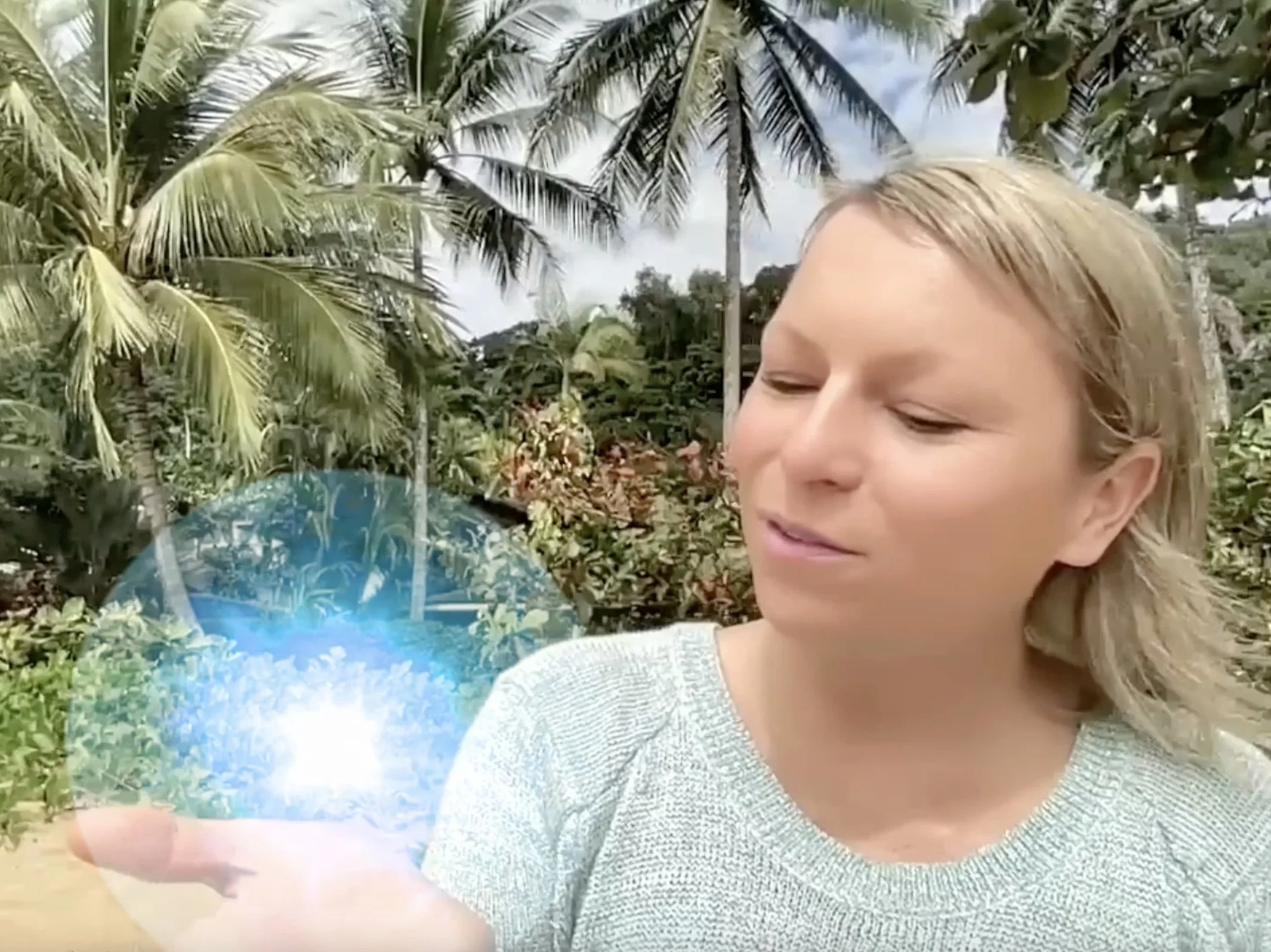 Julie Sulter is holding a glowing blue sphere in her hand, standing outdoors among tropical trees and palm trees.