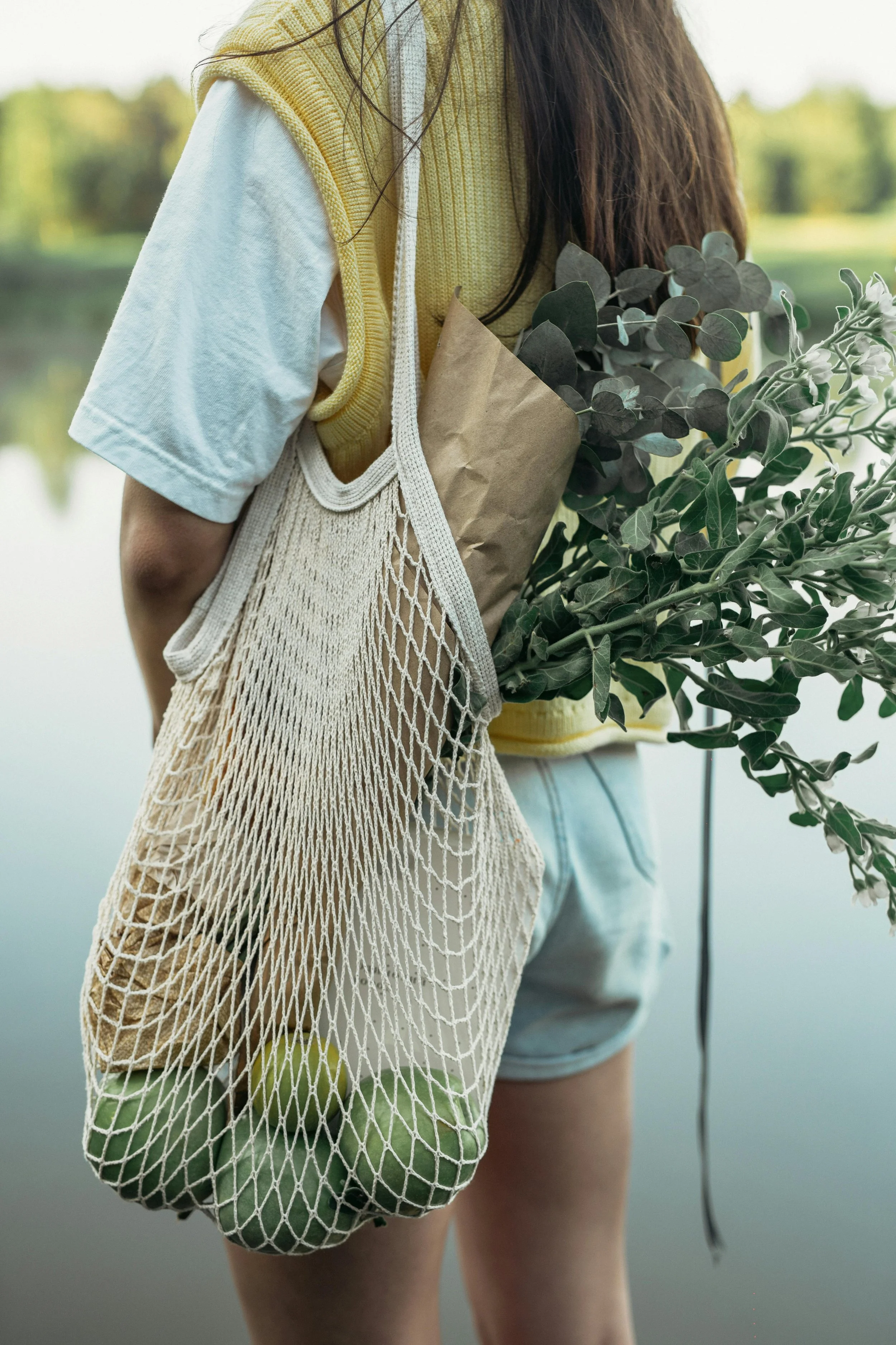 Person standing near a lake, holding a paper bag with eucalyptus and other greenery, wearing a yellow vest, white t-shirt, and denim shorts.