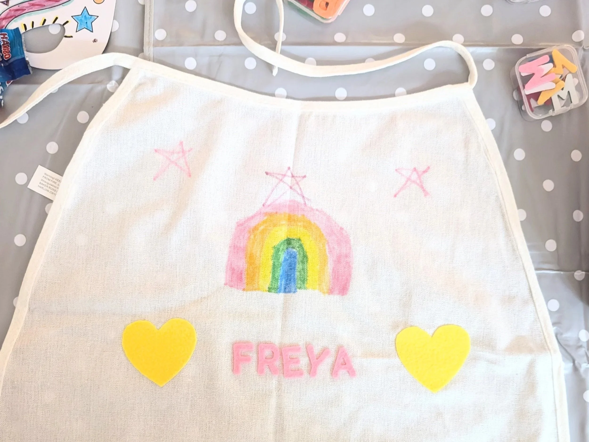 A fabric cape with a hand drawn rainbow and stars. Fabric letters and hearts have been stuck on during a craft party.