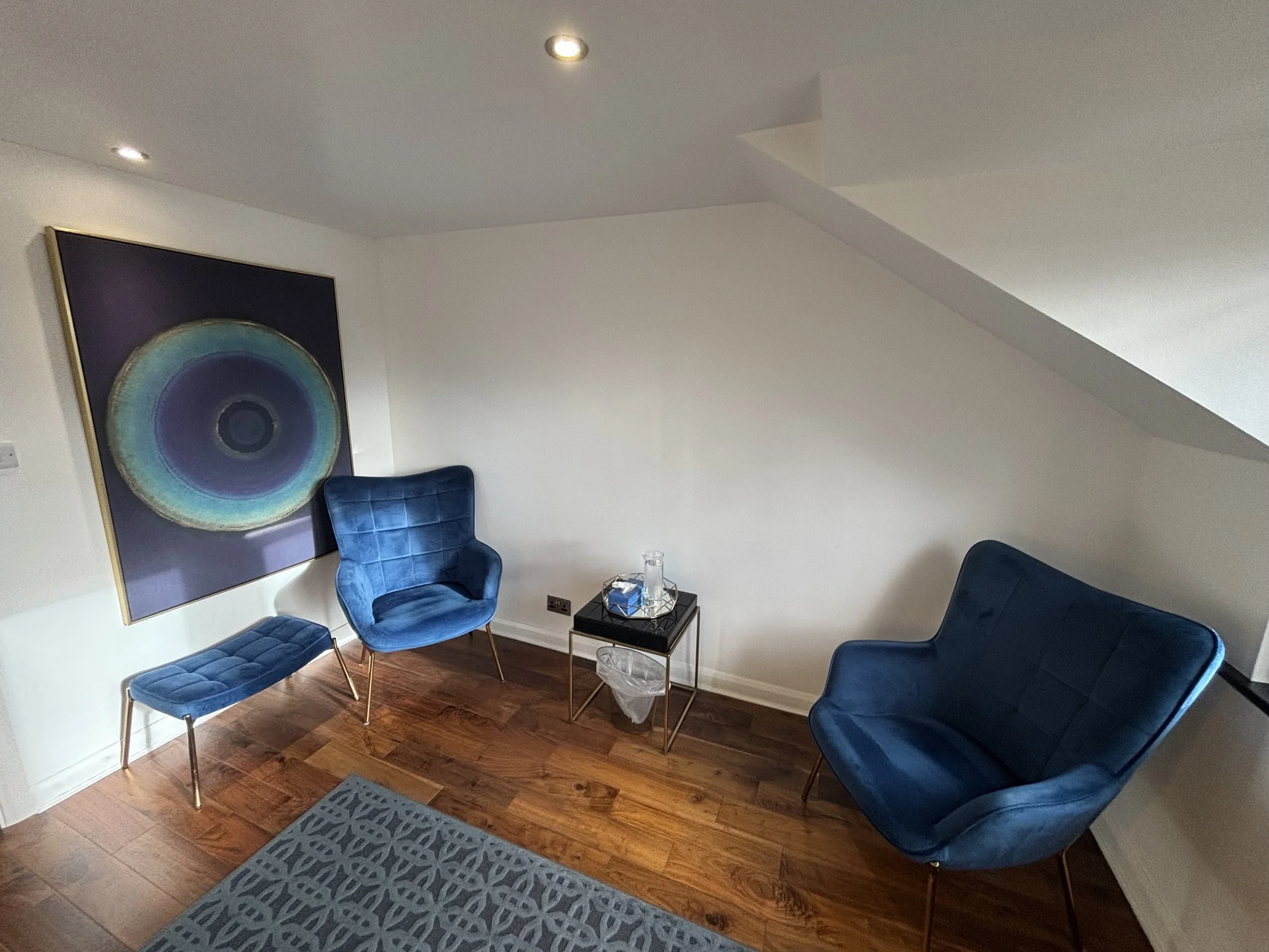 Living room corner with modern blue velvet armchair, matching blue velvet bench, and a small black side table with glassware. Large colorful circular abstract artwork on white wall, hardwood floor, and patterned gray rug.