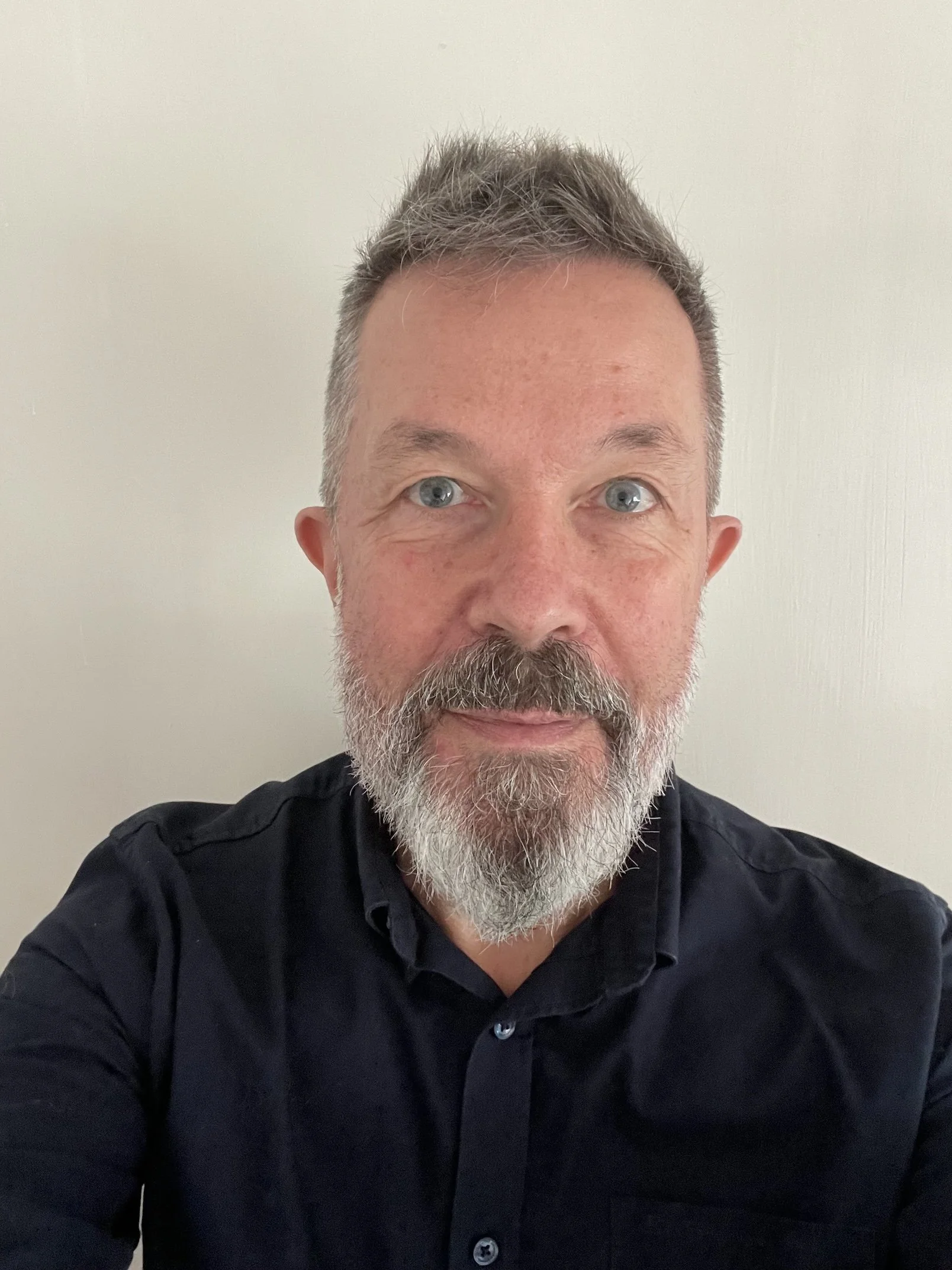 A middle-aged man with short gray hair, blue eyes, and a gray beard, wearing a black button-up shirt, standing against a plain white wall.