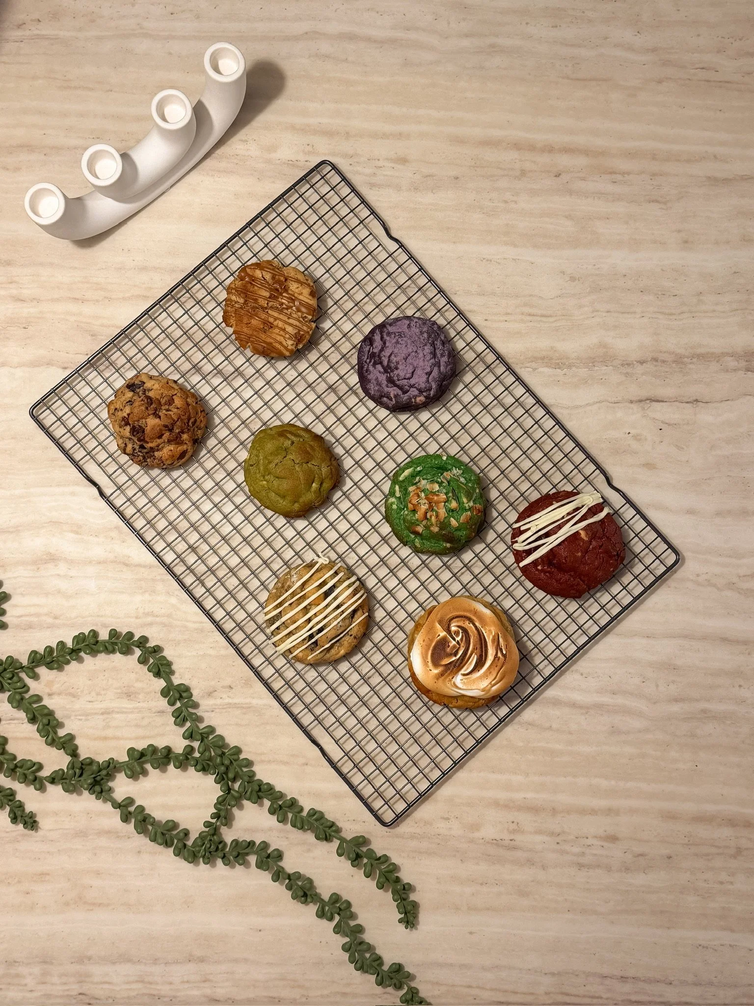 A metal cooling rack with decorated cookies on a light wooden surface. There is a white candlestick holder in the top left corner and a green vine on the bottom left.