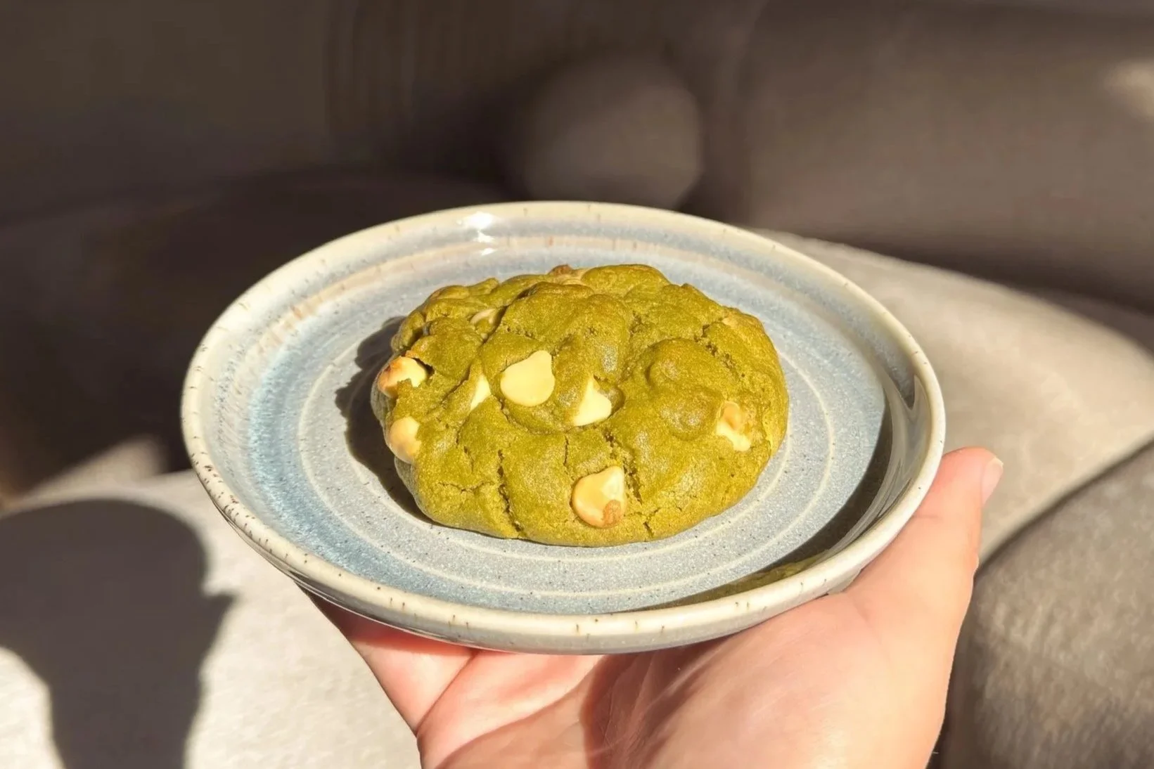 A person holding a small ceramic plate with a green matcha-flavored cookie containing white chocolate chips. The background is slightly blurred with sunlight.