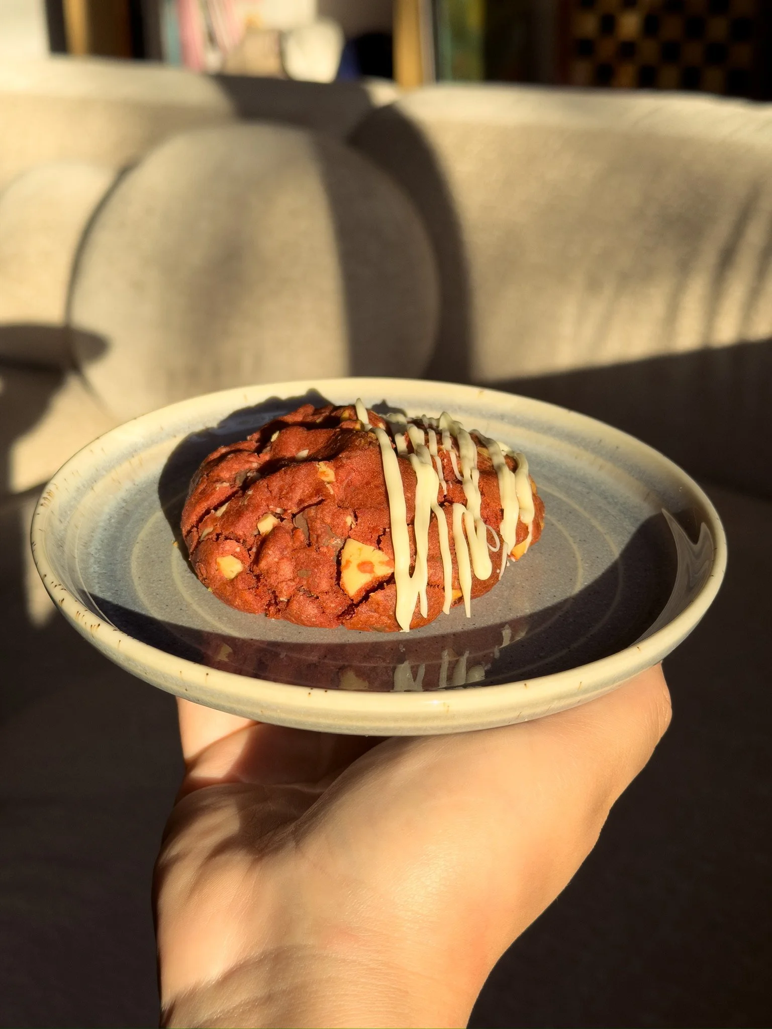 A hand holding a small bowl of cookie dough with white chocolate drizzle on top, in sunlight.