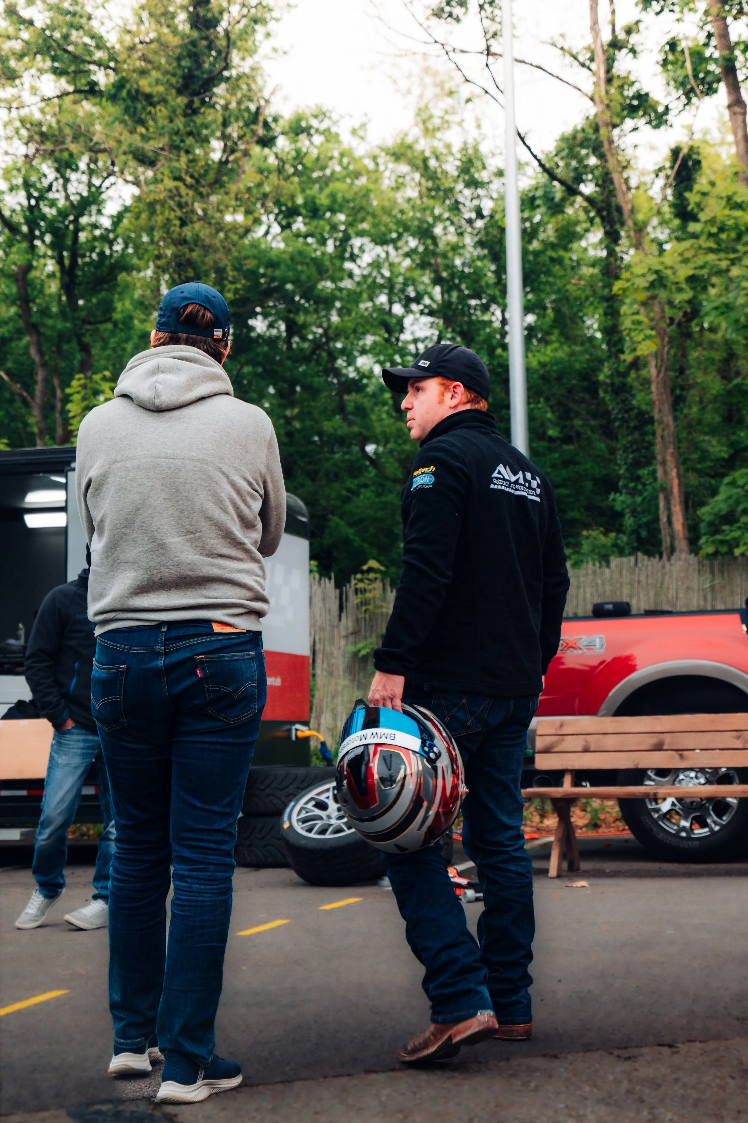 Zwei Männer im Gespräch bei einem Motorsport-Event, einer hält einen Helm, im Hintergrund ein Fahrzeuge und Reifen.
