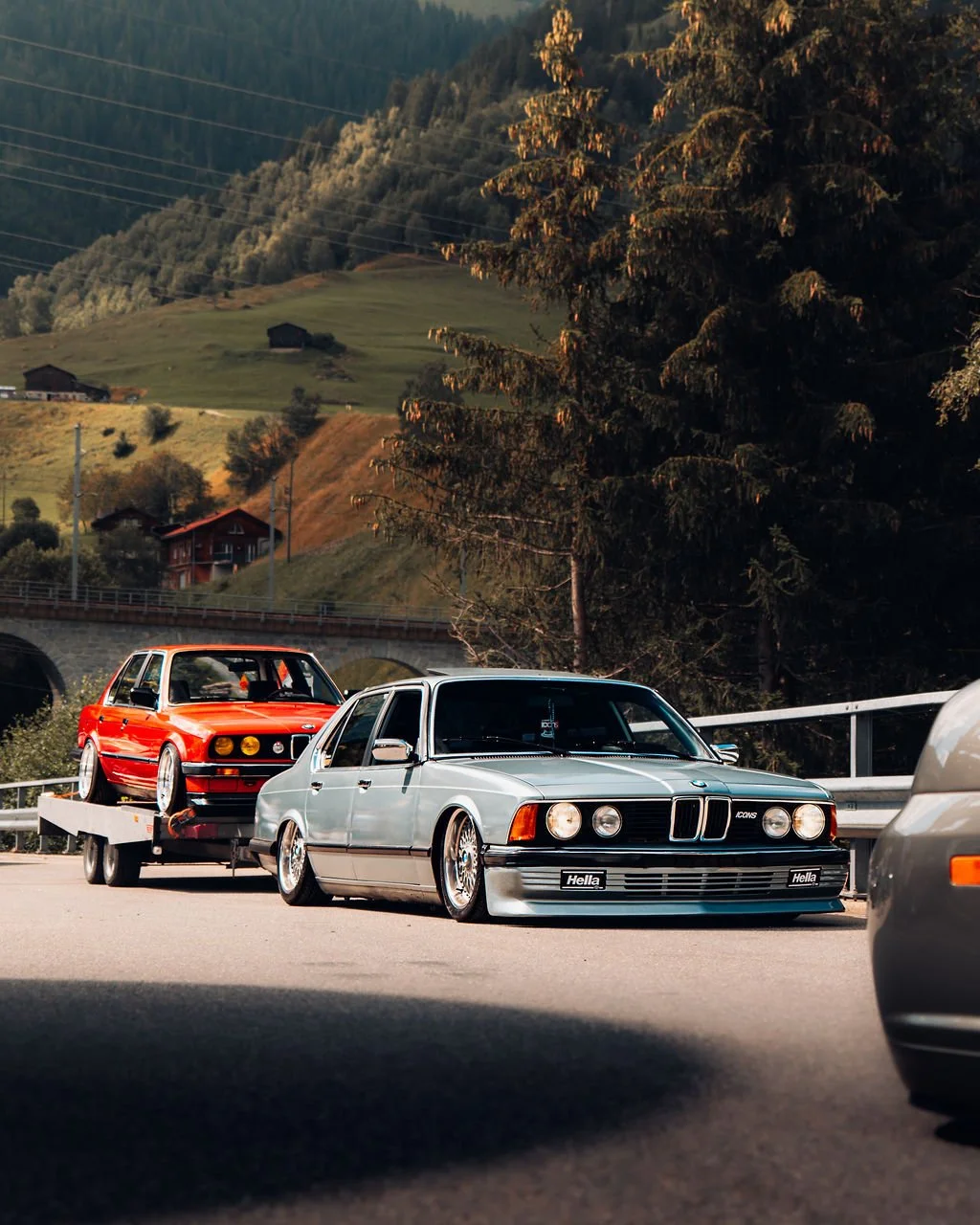 Ein silberner BMW E24 mit Tieferlegung und Classic Design fährt auf der Autobahn, hinter ihm ein roter BMW E30 auf einem Anhänger, umgeben von Bäumen und Hügeln in einer ländlichen Gegend.