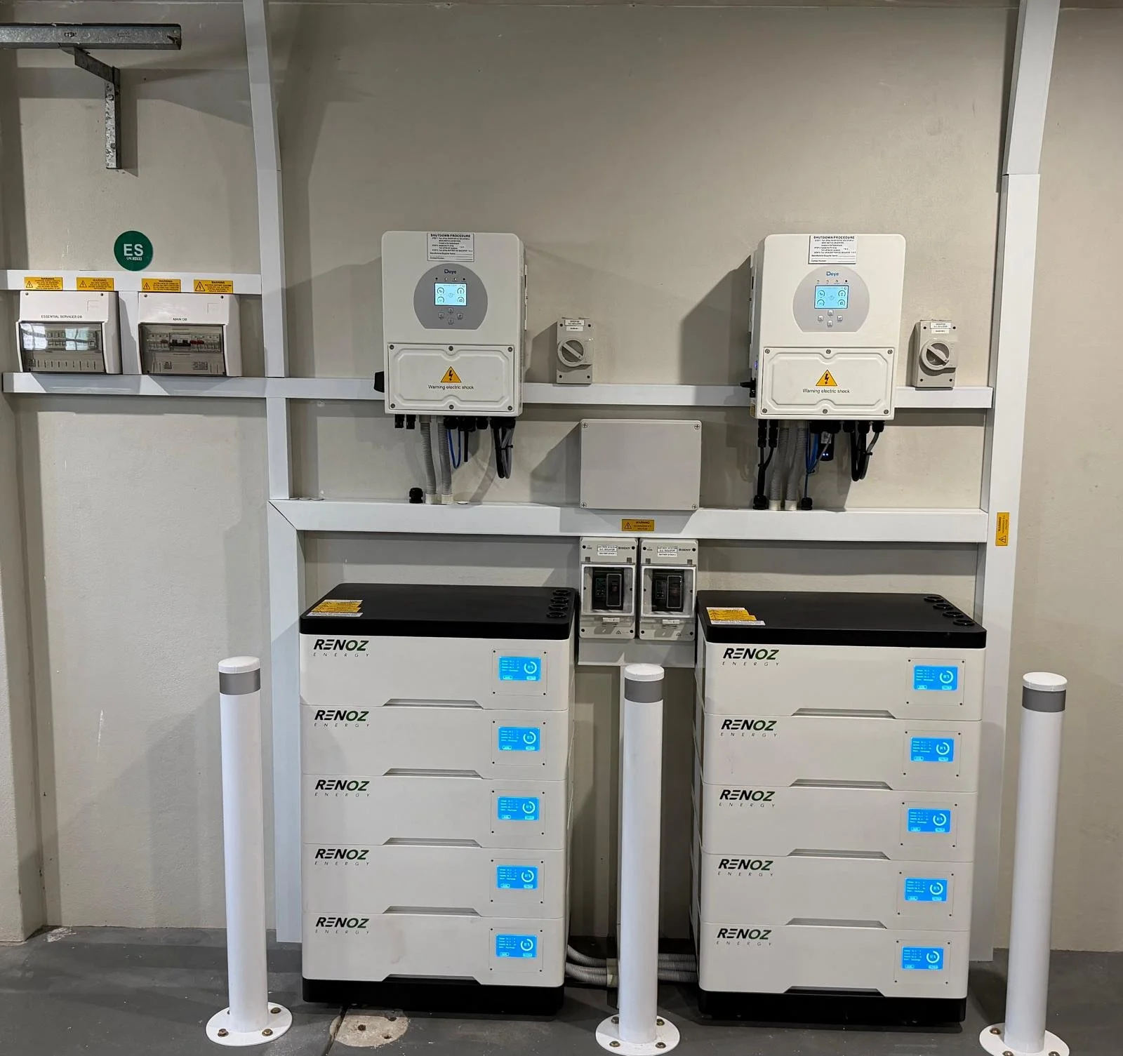 Two white RenoZ Energy battery storage units with digital displays and multiple compartments, positioned side by side, with electrical panels and meters mounted on a wall behind them, protected by white posts.