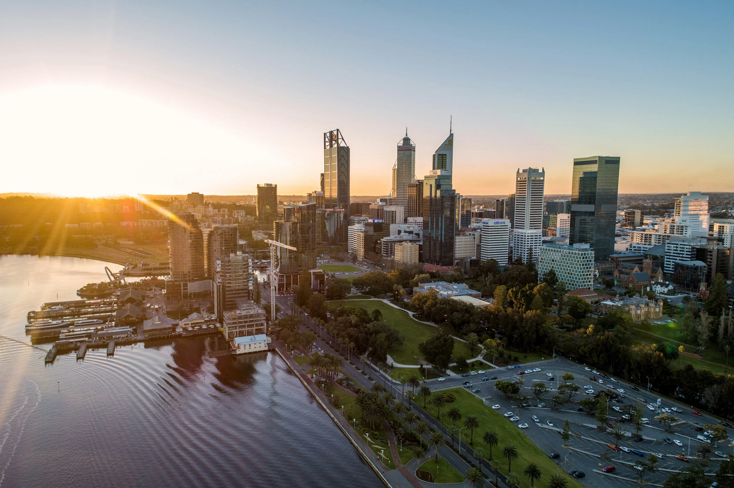 Aerial view of a city skyline at sunset, with tall skyscrapers, a river, waterfront buildings, and a park with trees and parking lot.