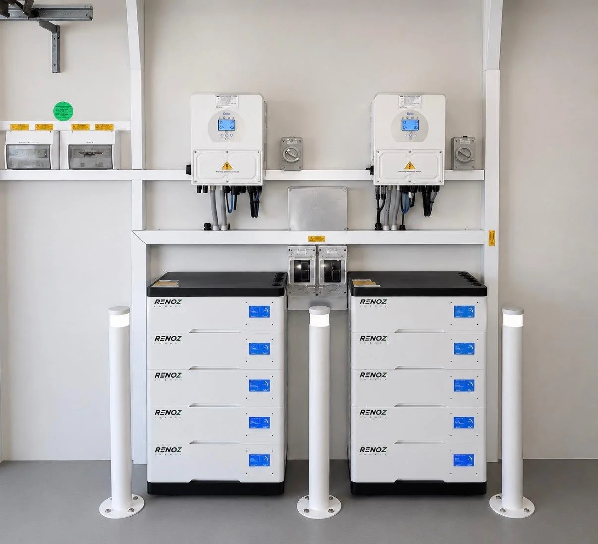 Two white RenoZ Energy battery storage units with digital displays and multiple compartments, positioned side by side, with electrical panels and meters mounted on a wall behind them, protected by white posts.