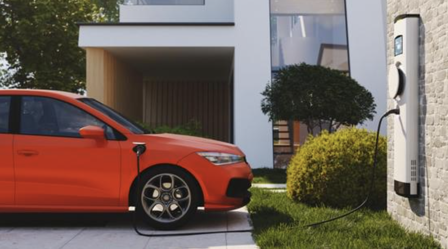 A red electric car is charging at a home charging station on a driveway, with a modern house and landscaped yard in the background.