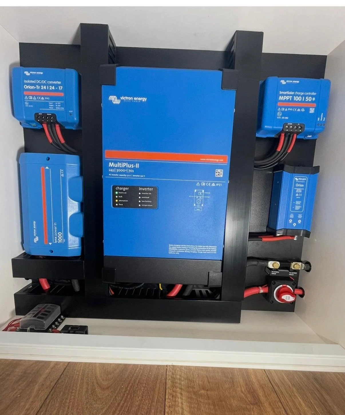 An electrical power inverter and related components mounted on a black frame inside a small enclosure. The setup includes blue boxes labeled with brands like Victron Energy, with various connecting cables.
